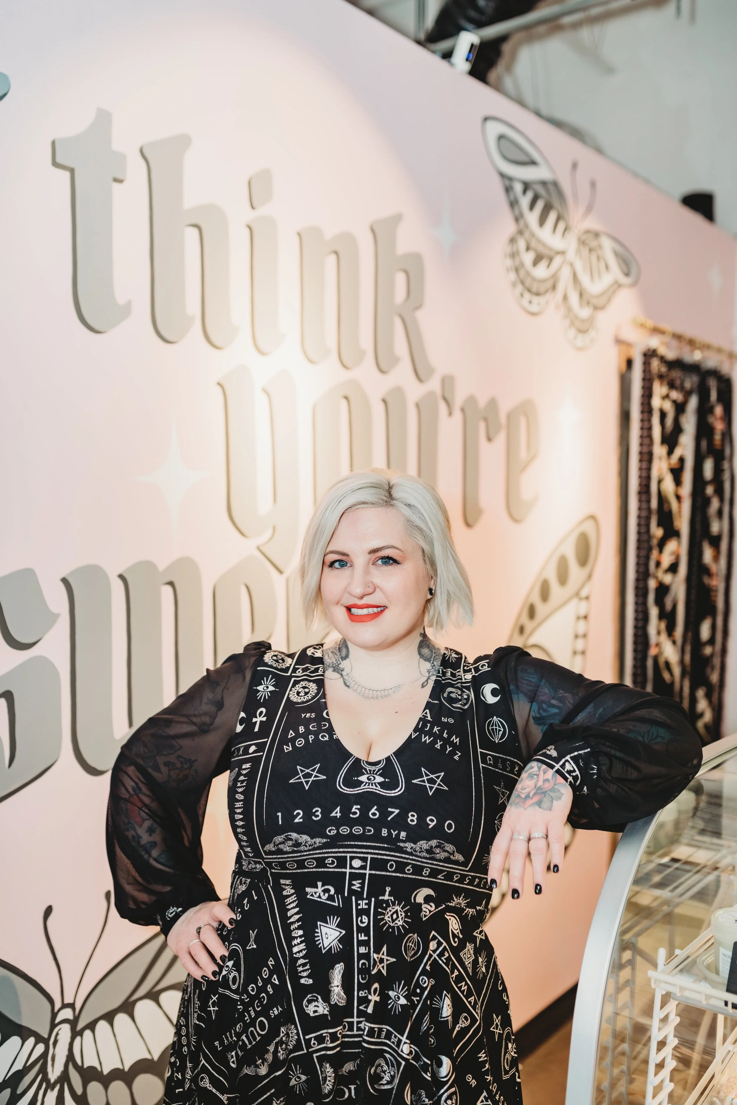 Person with blonde hair, wearing a black dress with mystical symbols, stands in front of a pink wall with the phrase "I think you're sweet" and butterfly illustrations.