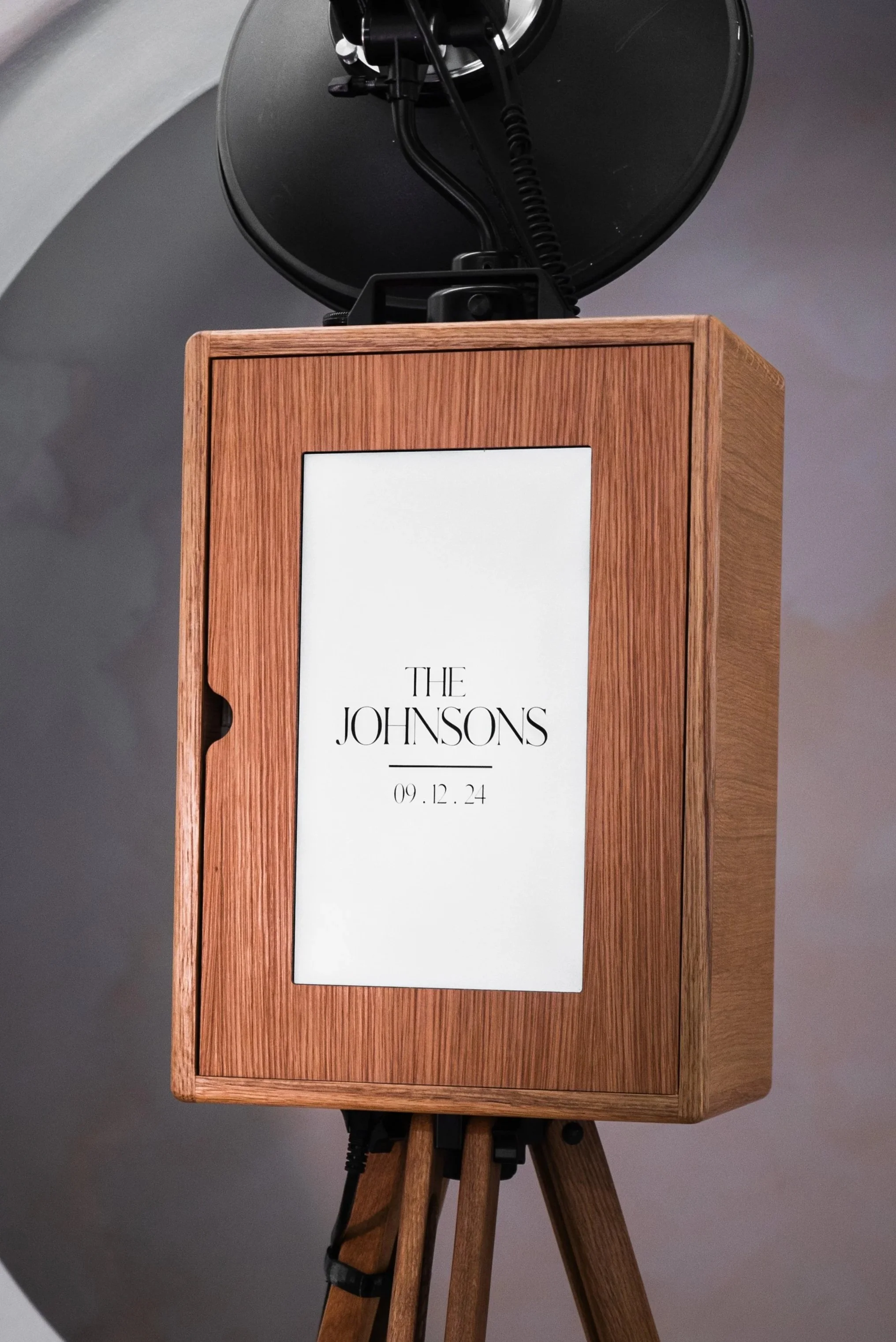 A wooden photo booth with a screen displaying the text 'The Johnsons' and a date '09.12.24,' standing on a wooden tripod.