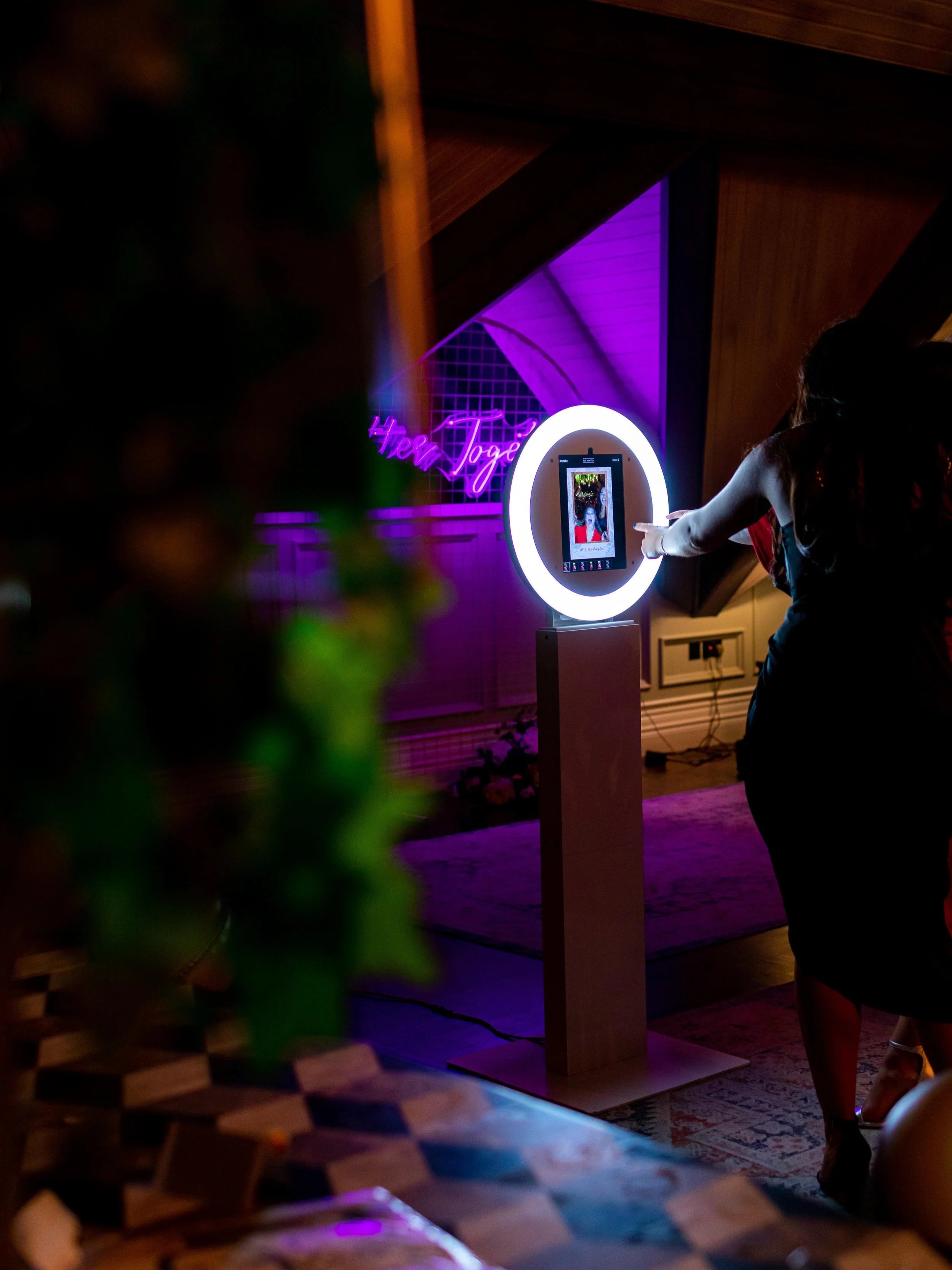 A woman using a photo booth with a circular ring light in a dimly lit room with purple lighting. There is a neon sign in the background that says 'Here To'.