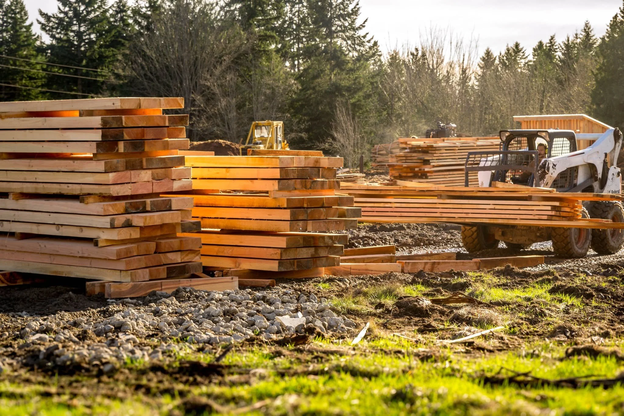 A skid steer places a stack of lumber onto a larger pile of large wooden boards