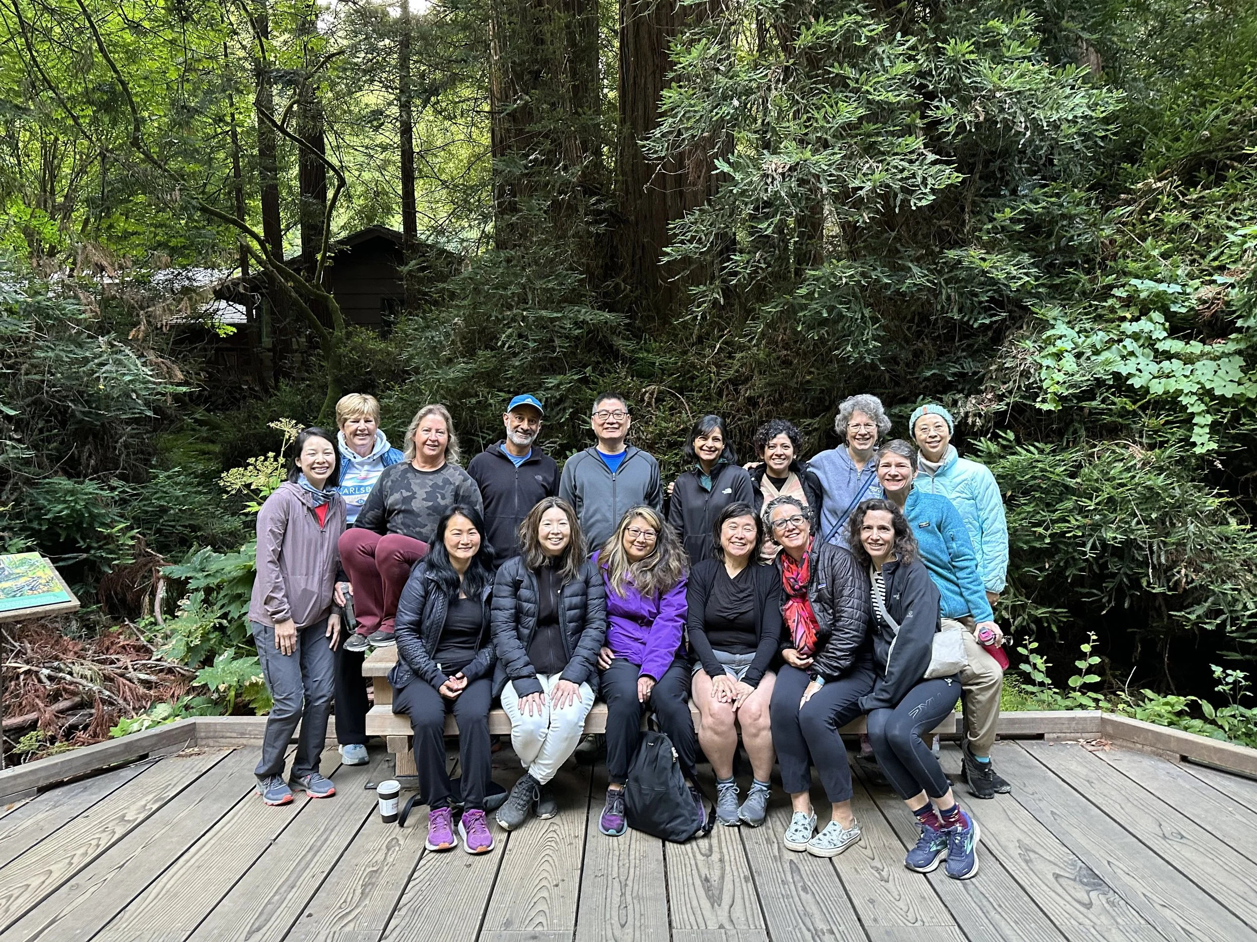 A group of fifteen people, mostly women, posing together on a wooden platform in a lush forest with tall trees and greenery in the background.