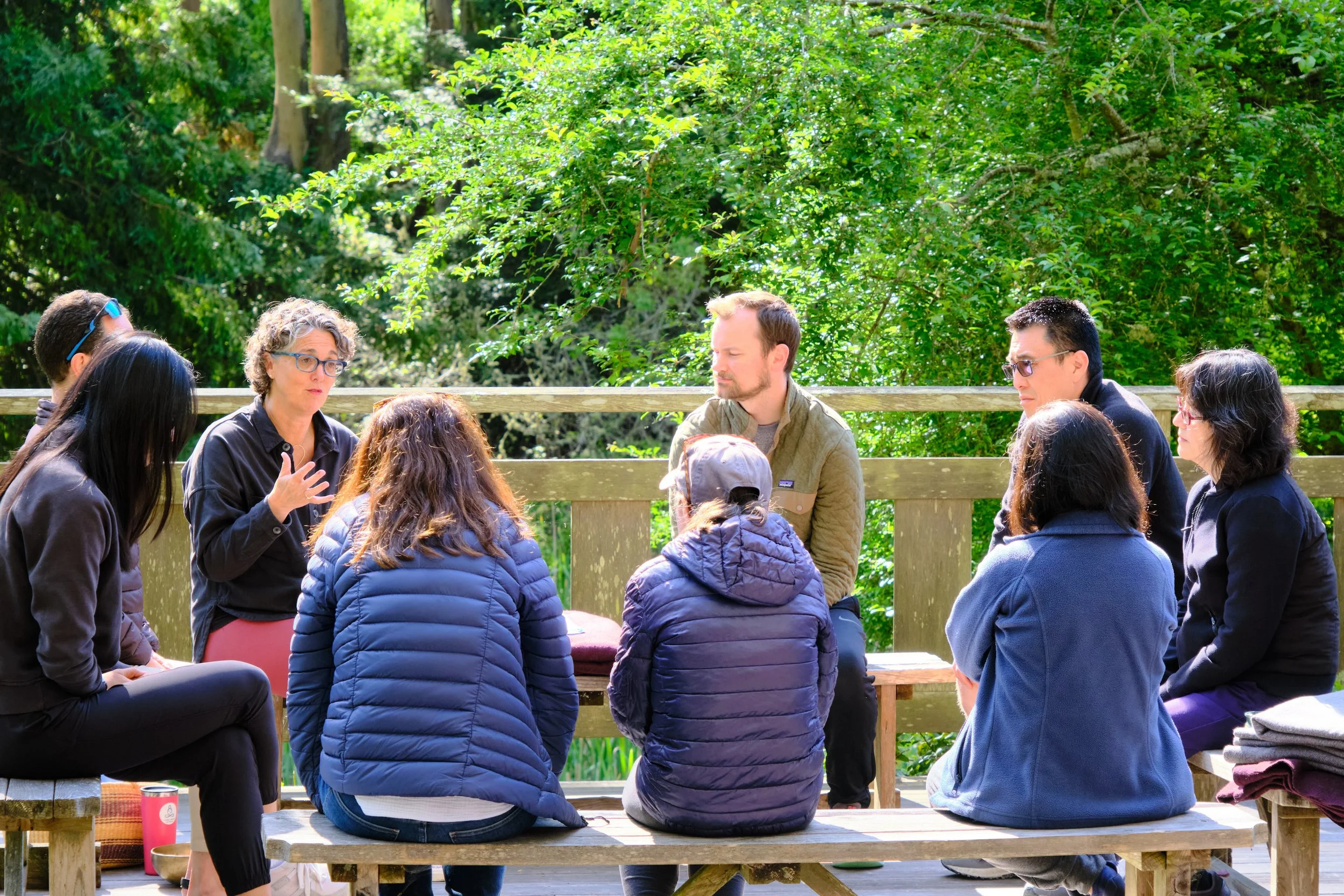 A group of eight people sitting outdoors in a circle, engaged in a discussion or meeting, surrounded by green trees and foliage.