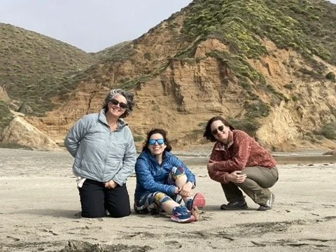 Three women on a beach with hills in the background. One woman is standing, and two are squatting, all smiling at the camera.