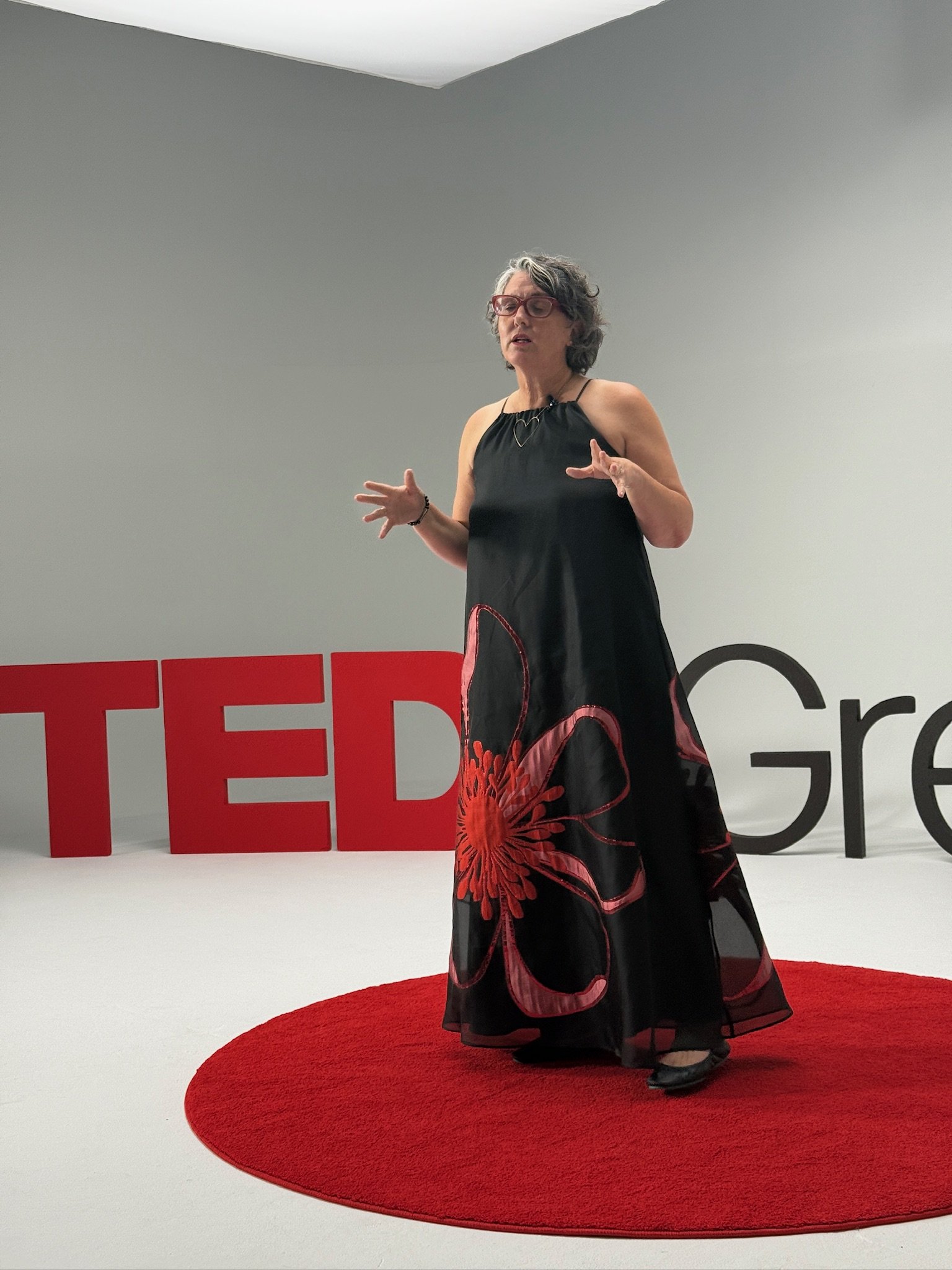 A woman standing on a red circular carpet, speaking in front of large red and black TEDx letters on a plain background.