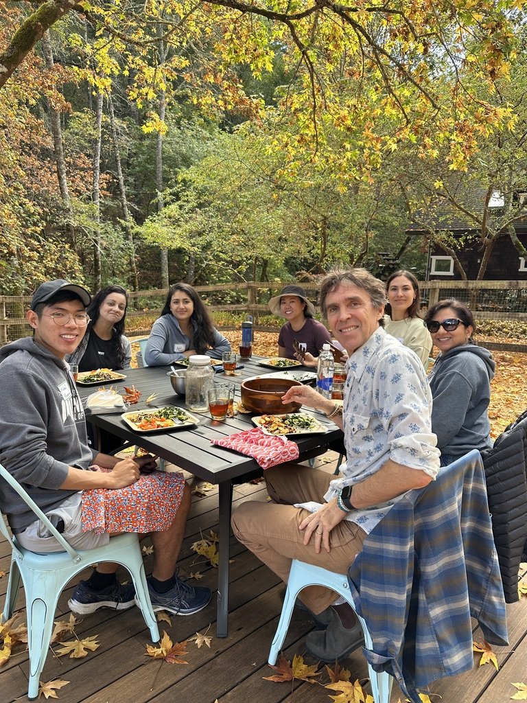 A group of seven people seated around a table outdoors in a wooded area during fall, enjoying a meal together.