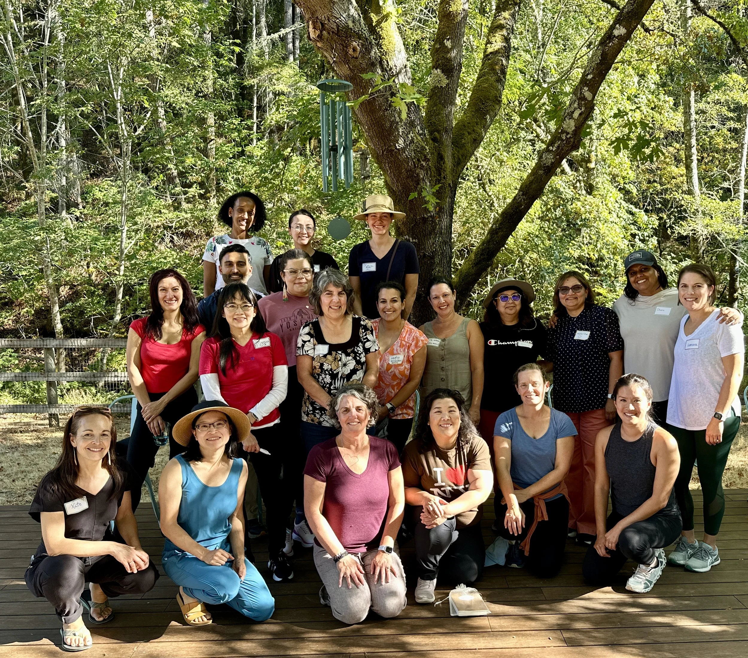 A group of 20 smiling women posing outdoors on a wooden deck in front of a large tree with green foliage, some wearing hats and casual clothing.