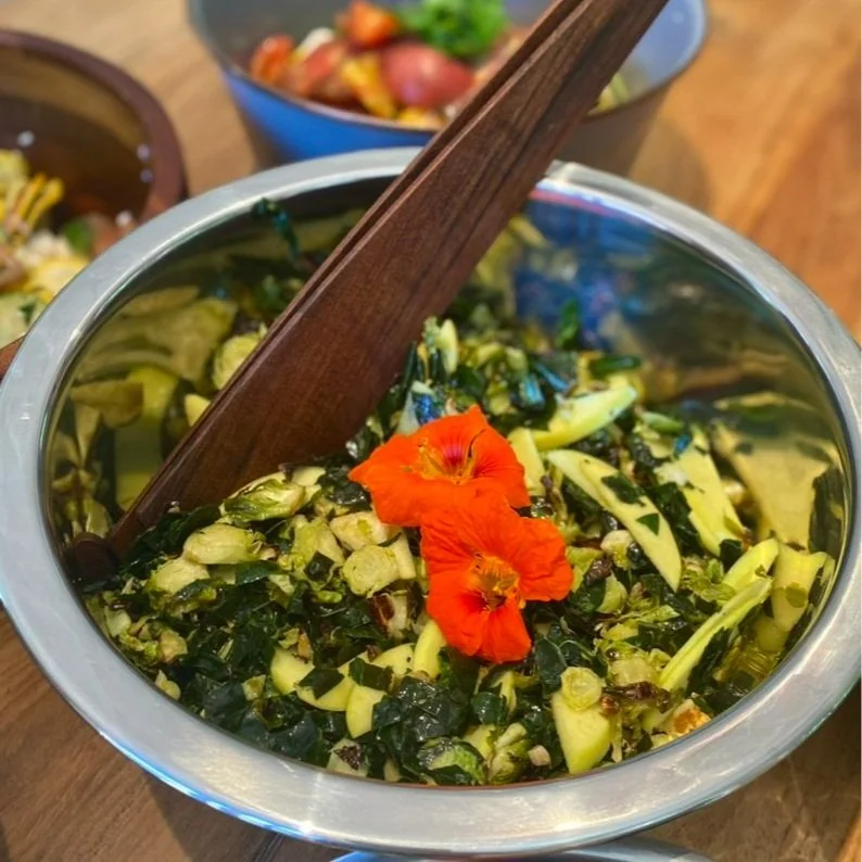 A metal bowl of mixed greens with edible orange flowers on top and a wooden salad fork inside, on a wooden table.
