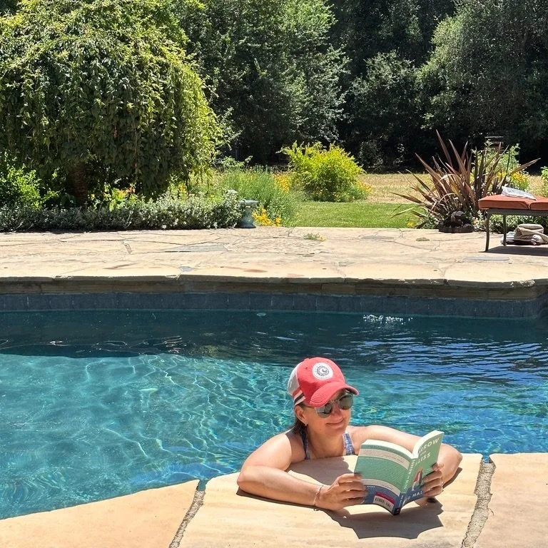 A woman in sunglasses and a baseball cap reading a book while leaning on the edge of a swimming pool in a backyard surrounded by trees and greenery.