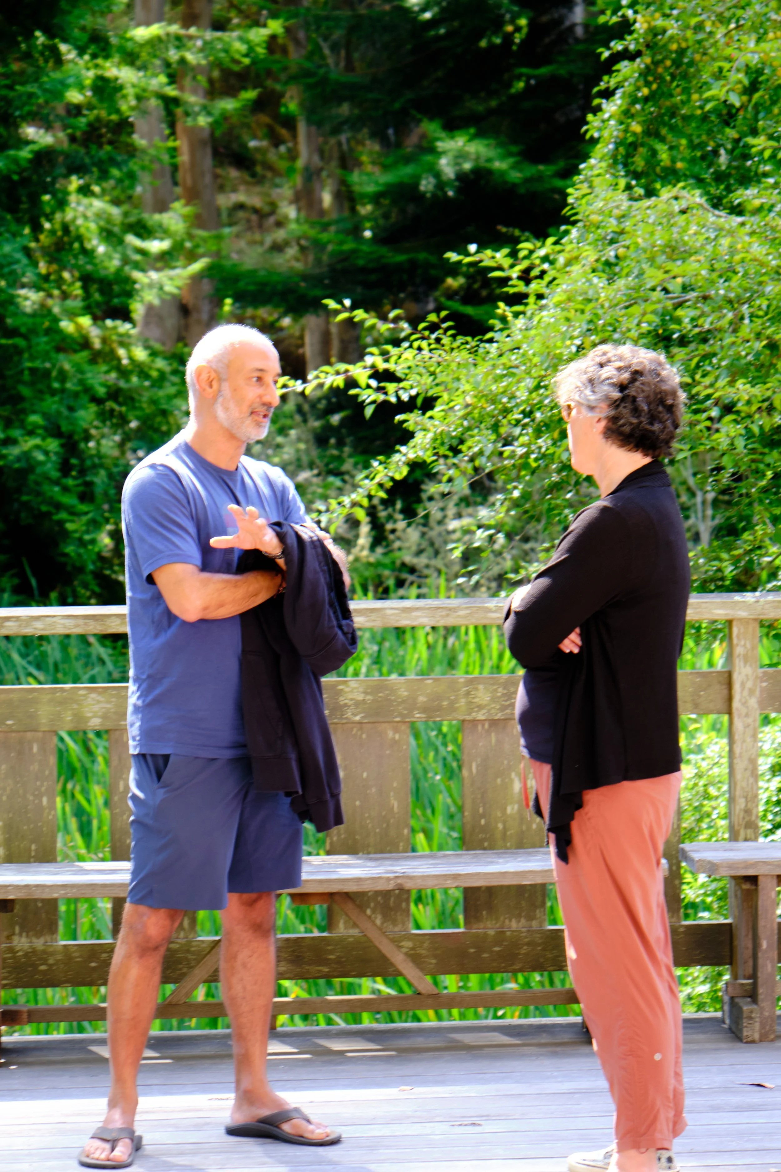 An older man and woman standing on a wooden deck, engaged in conversation, with lush green trees and foliage in the background.