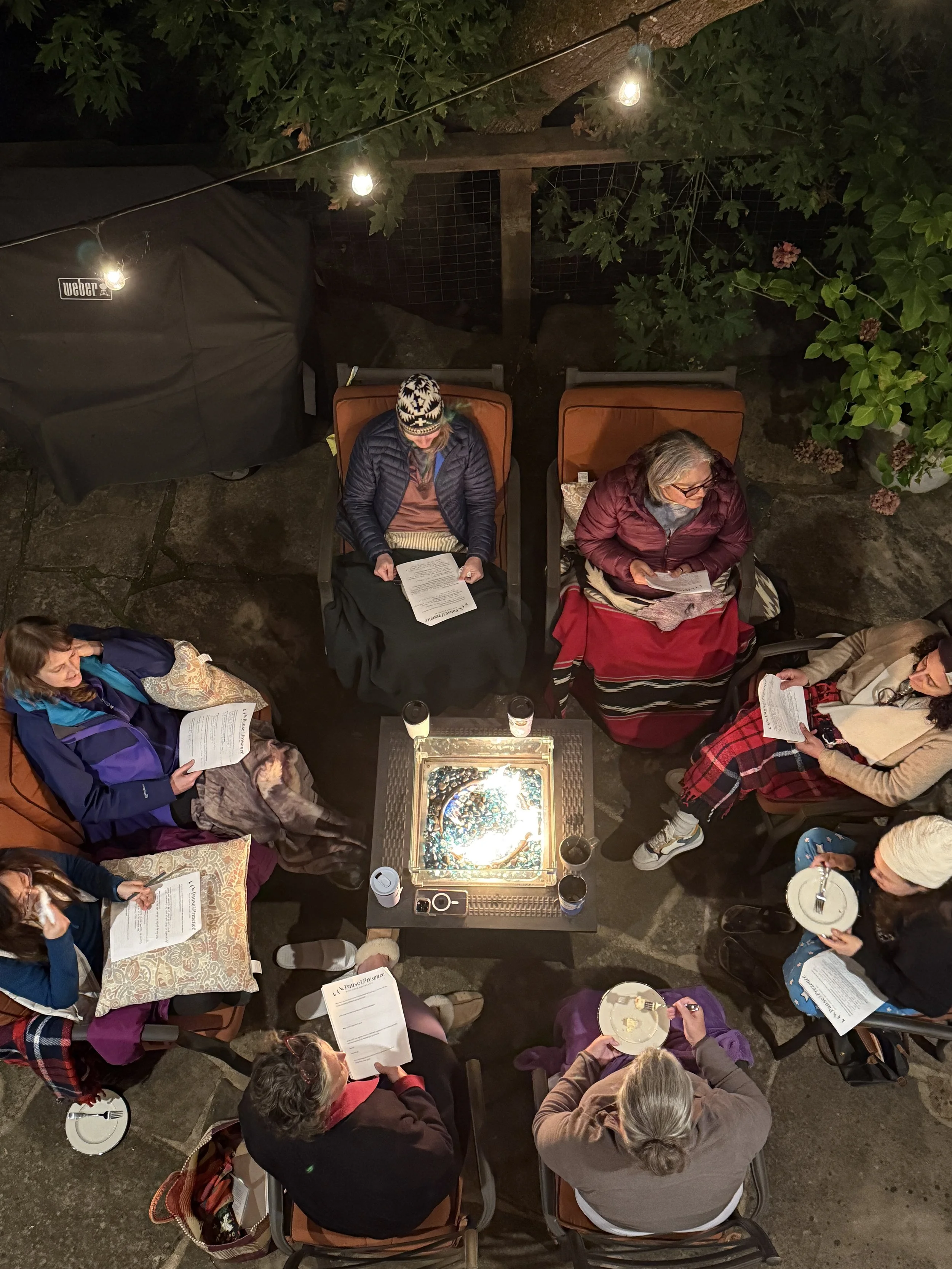 Group of people sitting around a square table outdoors at night, reading papers, with a fire pit in the center and string lights overhead.