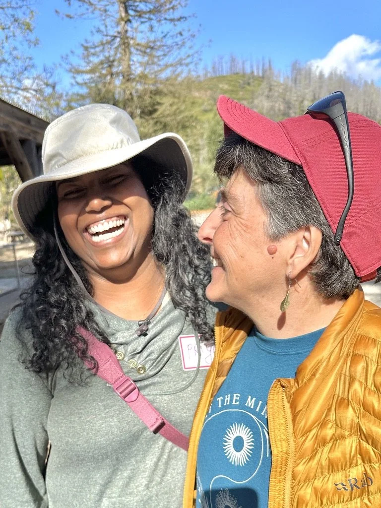 Two women outdoors, smiling and laughing at each other, with trees, blue sky, and a hill in the background.