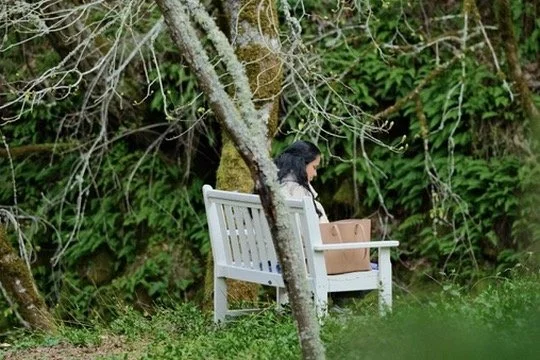 A woman sitting on a white bench outdoors in a wooded area.