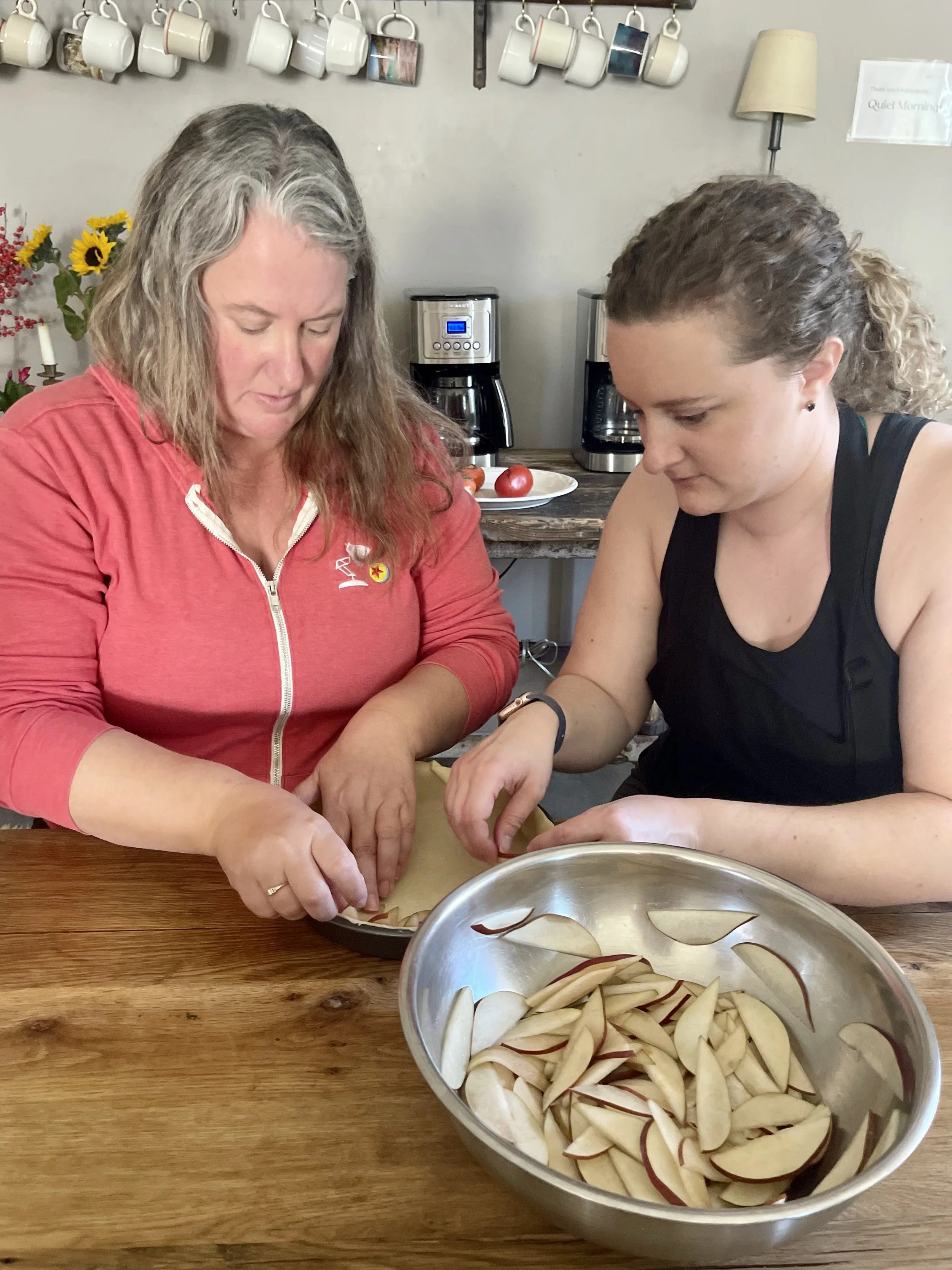 Two women preparing sliced apples in a kitchen, with a metal bowl of apple slices on a wooden table. They are working together, possibly making a pie or other dish.