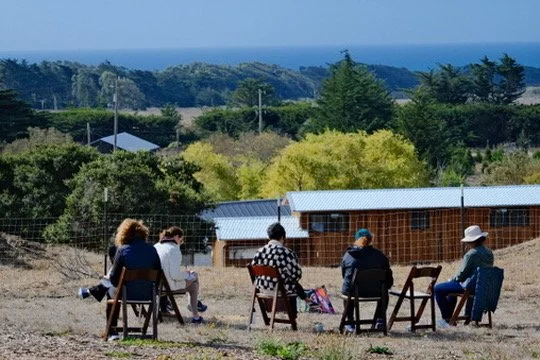 Five people sitting on chairs outdoors, facing a scenic landscape with trees and a body of water in the distance.