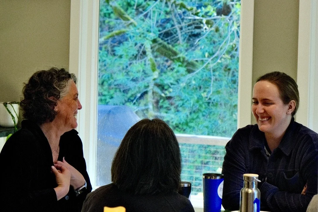 Three people sitting at a table inside, engaging in conversation and smiling, with a large window showing trees outside.
