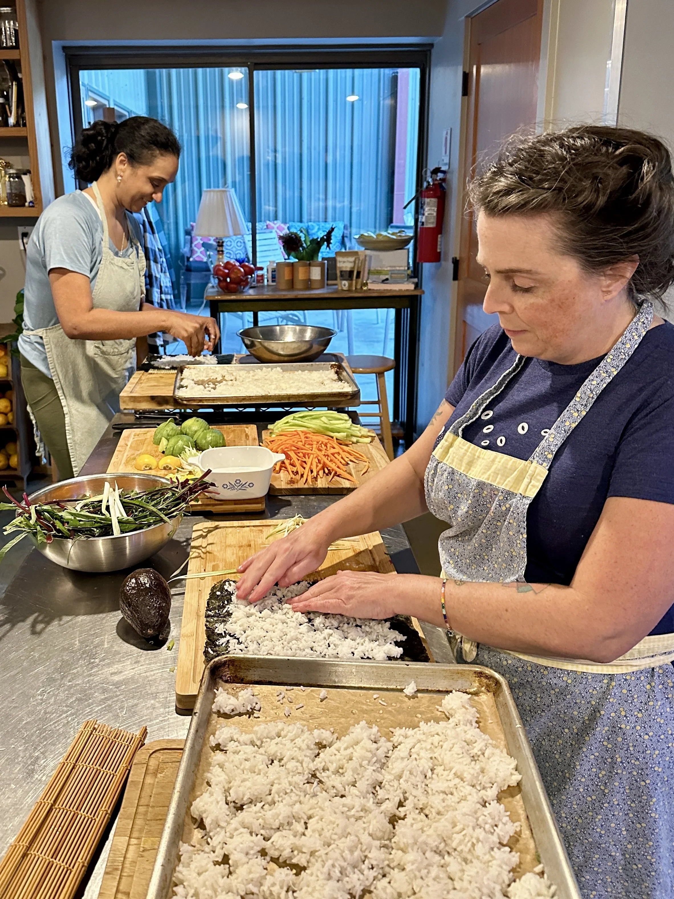 Two women preparing sushi in a kitchen, with rice, vegetables, and sushi rolling mats on the counter.