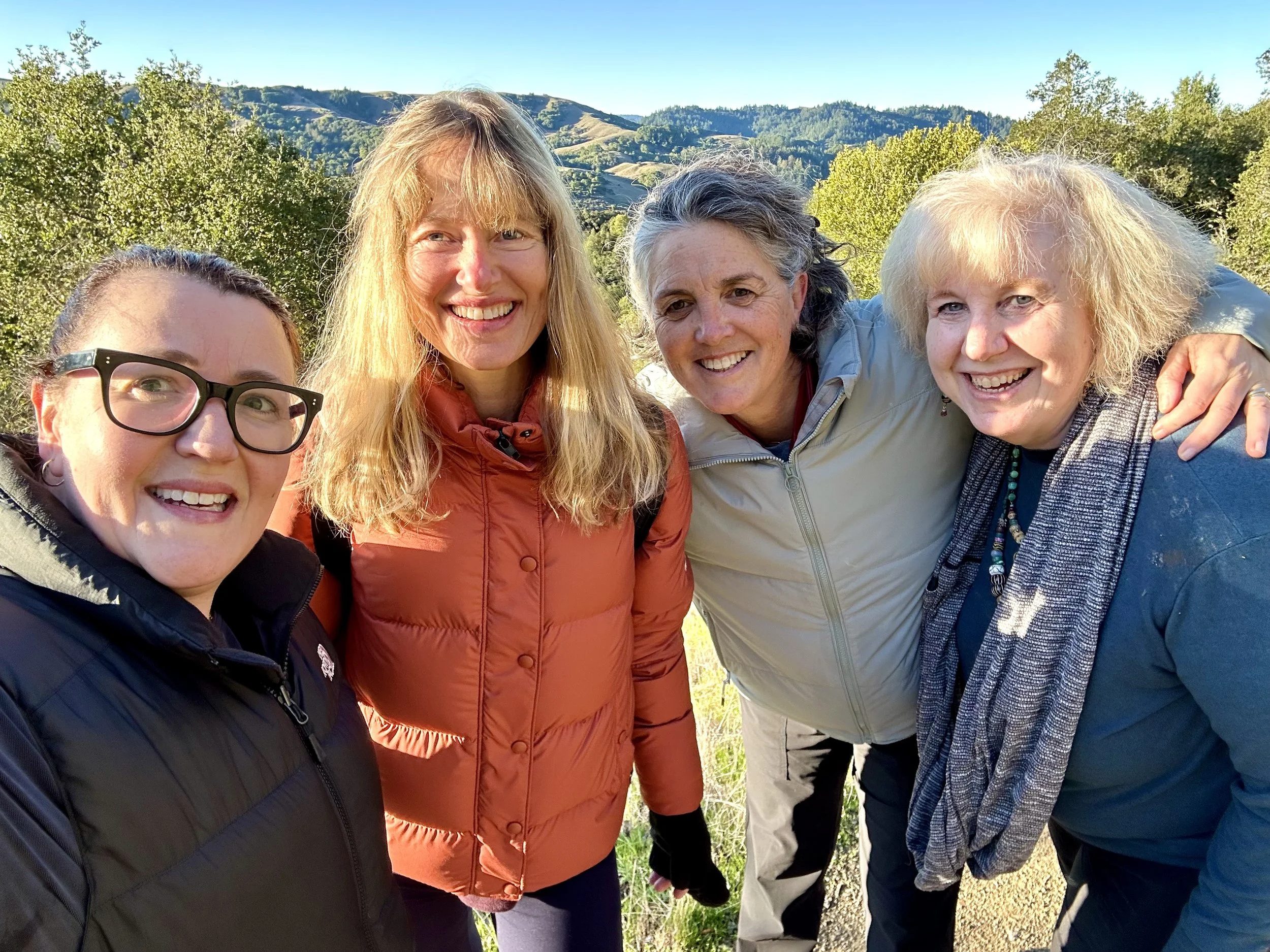 Four smiling women outdoors with hills and trees in the background.