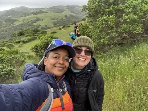 Two women hiking outdoors in a lush green landscape with rolling hills and trees, smiling at the camera.