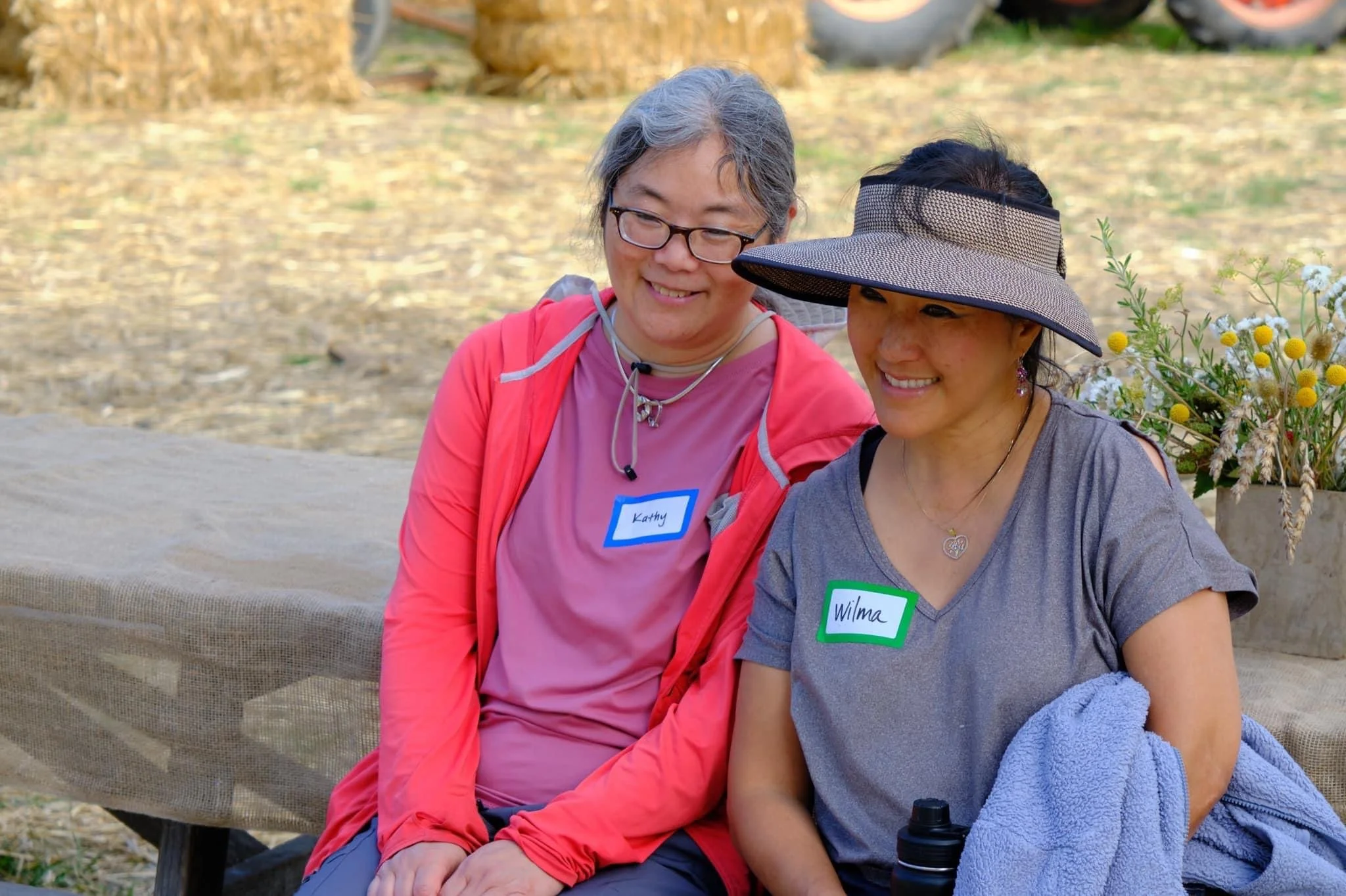 Two women sitting on a bench outdoors, smiling. One woman has gray hair, glasses, a pink shirt, and a name tag that reads 'Kathy.' The other woman has dark hair, a wide-brimmed hat, a gray T-shirt, and a name tag that reads 'Wilma.' There are flowers