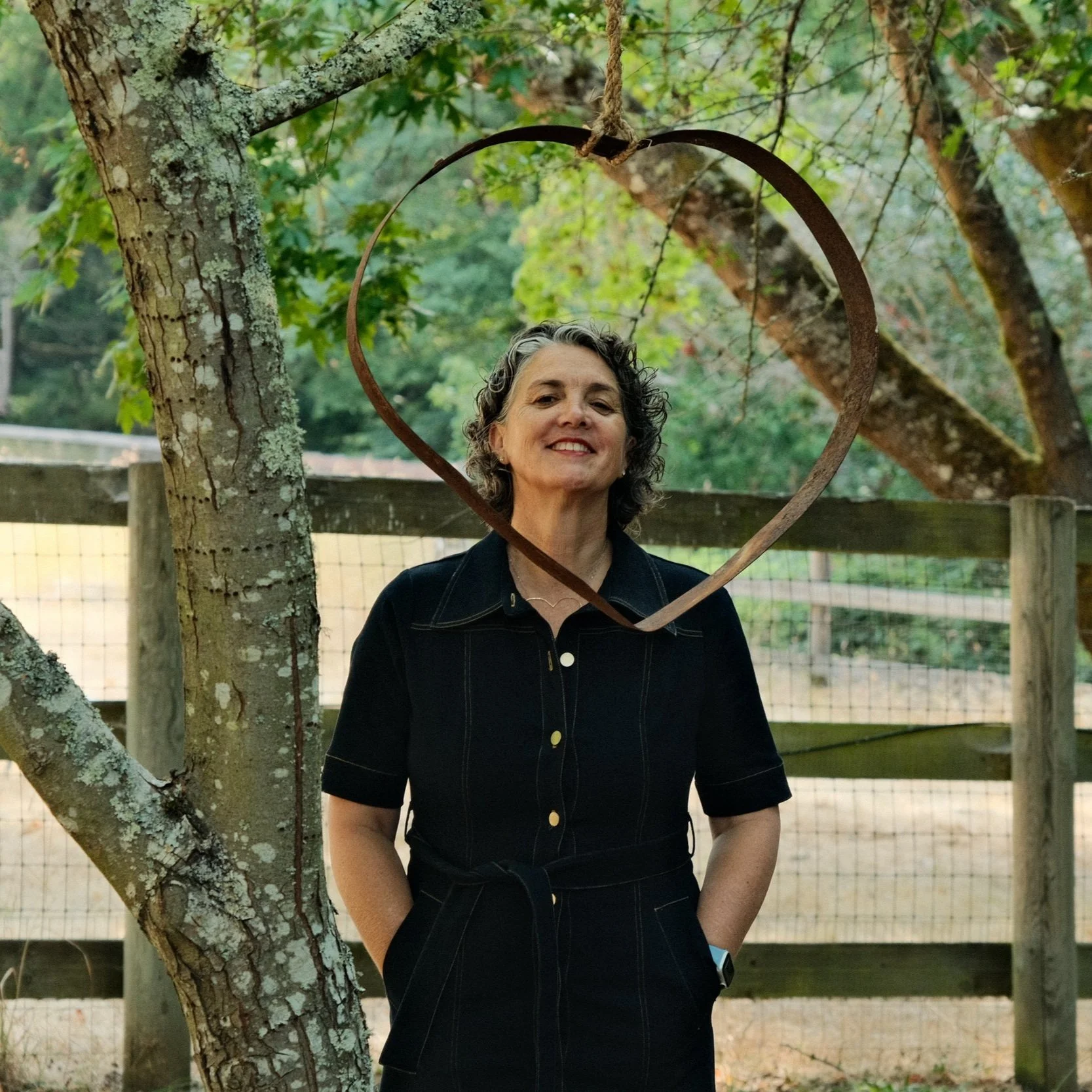 A woman with curly gray hair and a black dress standing outdoors in front of a tree with a heart-shaped metal frame hanging from a branch.