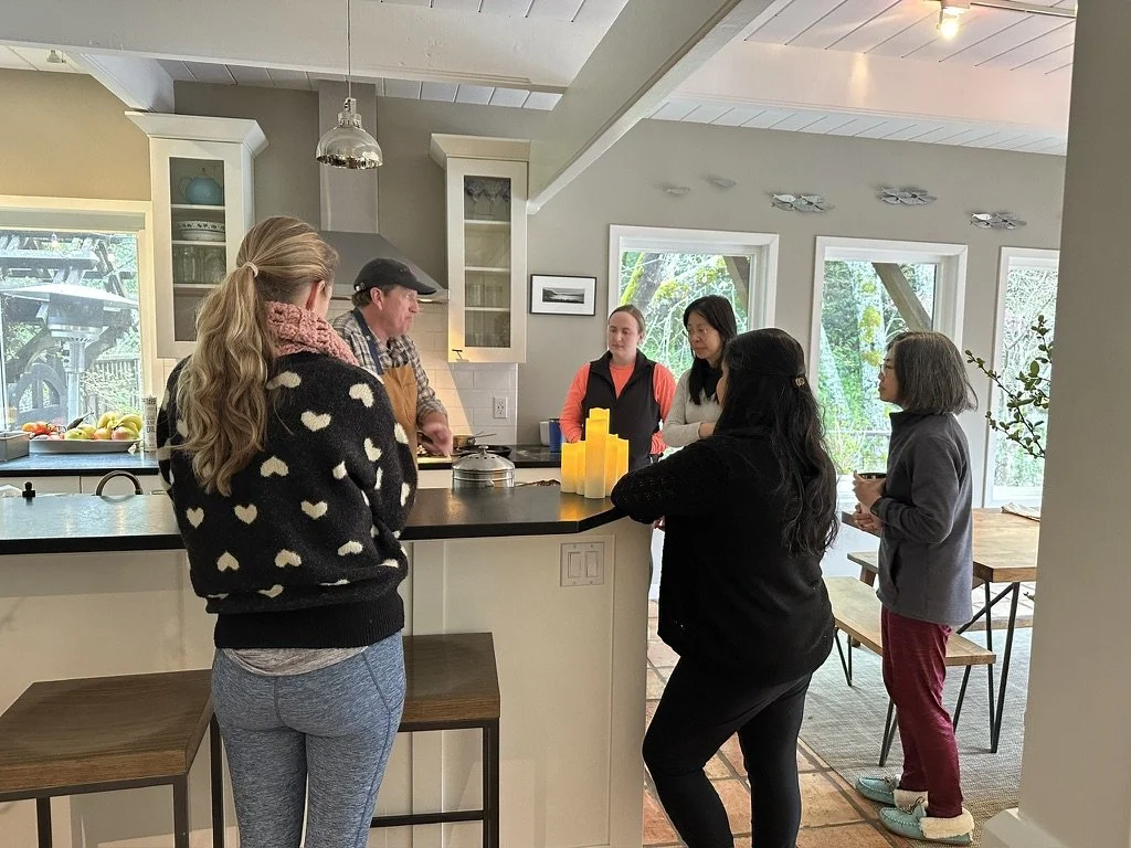 A group of people gathered in a kitchen, listening to a man who appears to be demonstrating something at the counter. The kitchen has white cabinets, a window with trees outside, and a dining table in the background.