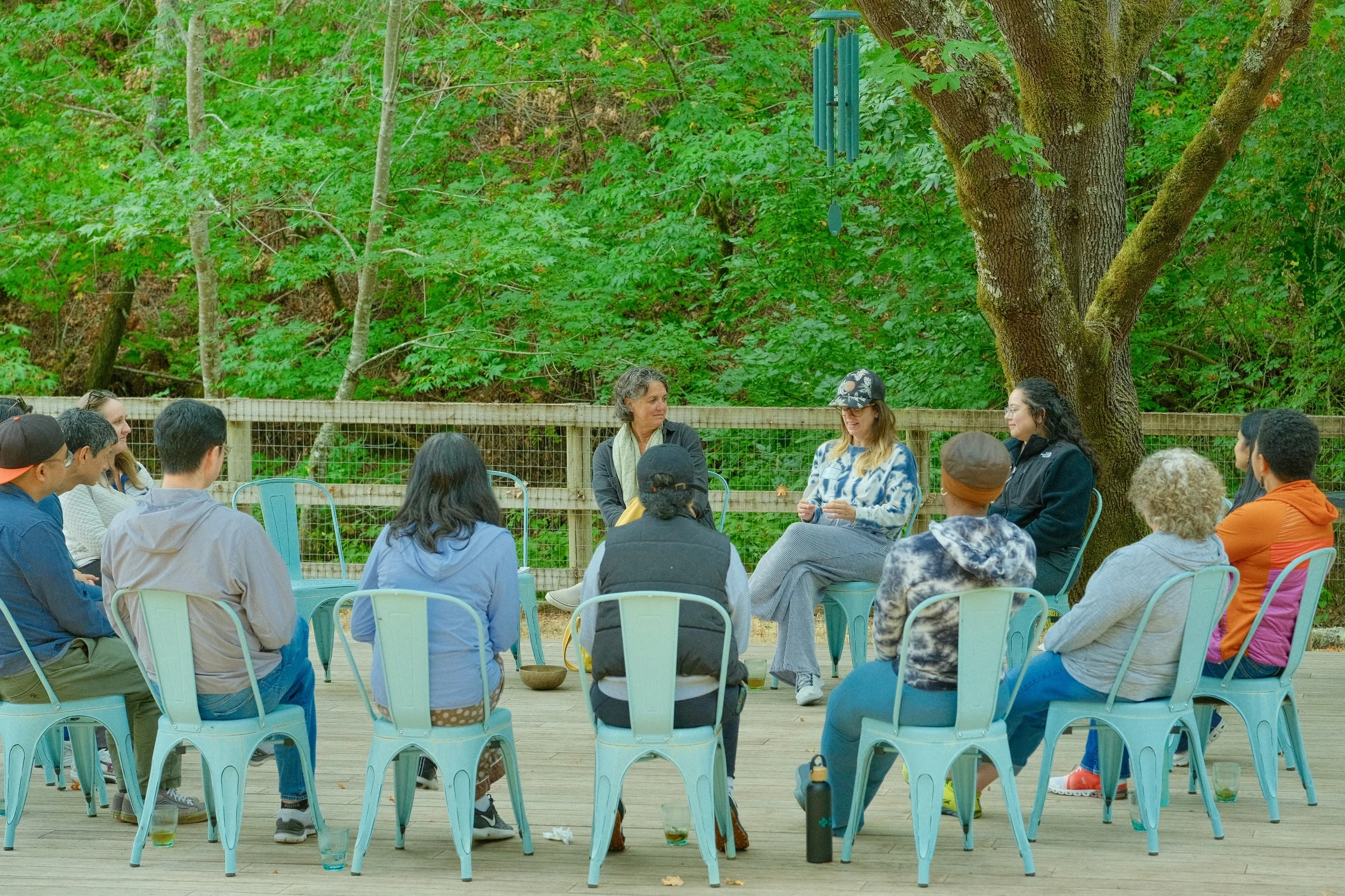 A group of people sitting on chairs in an outdoor setting with trees and greenery in the background, participating in a discussion or workshop.