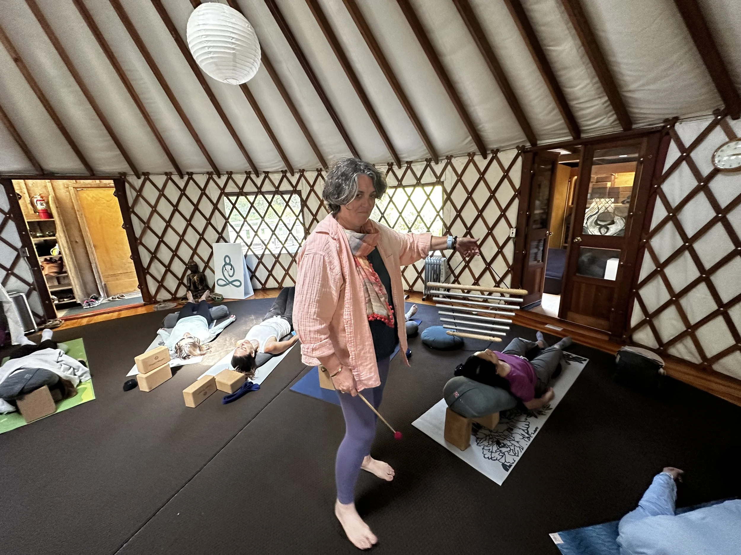 A woman in a yoga class pointing at students lying on mats with yoga blocks and bolsters in a yurt-style studio with wooden lattice walls and a lantern hanging from the ceiling.