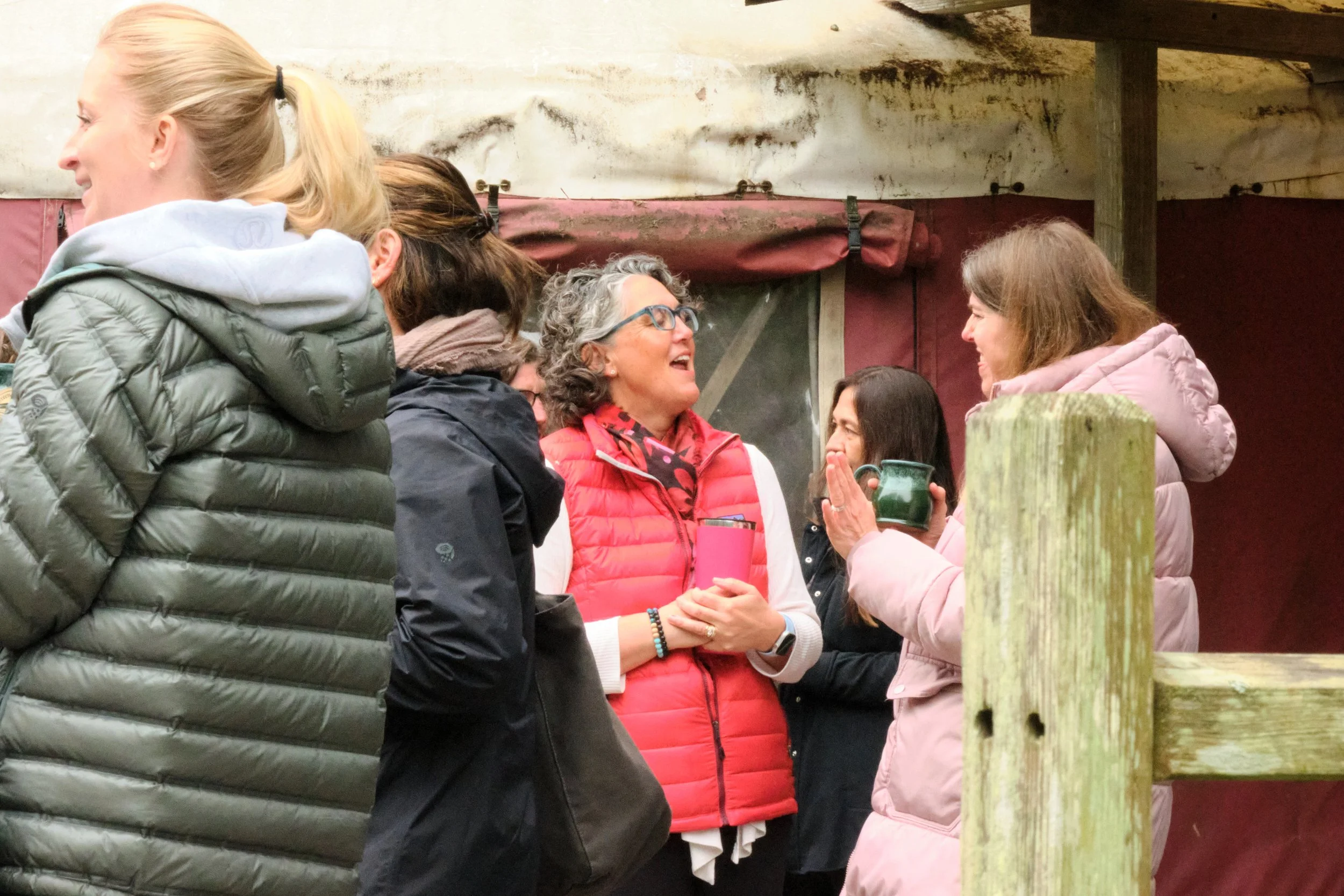 Group of women, mostly wearing puffer jackets, engaging in conversation outdoors near a wooden fence, with one woman holding a mug.