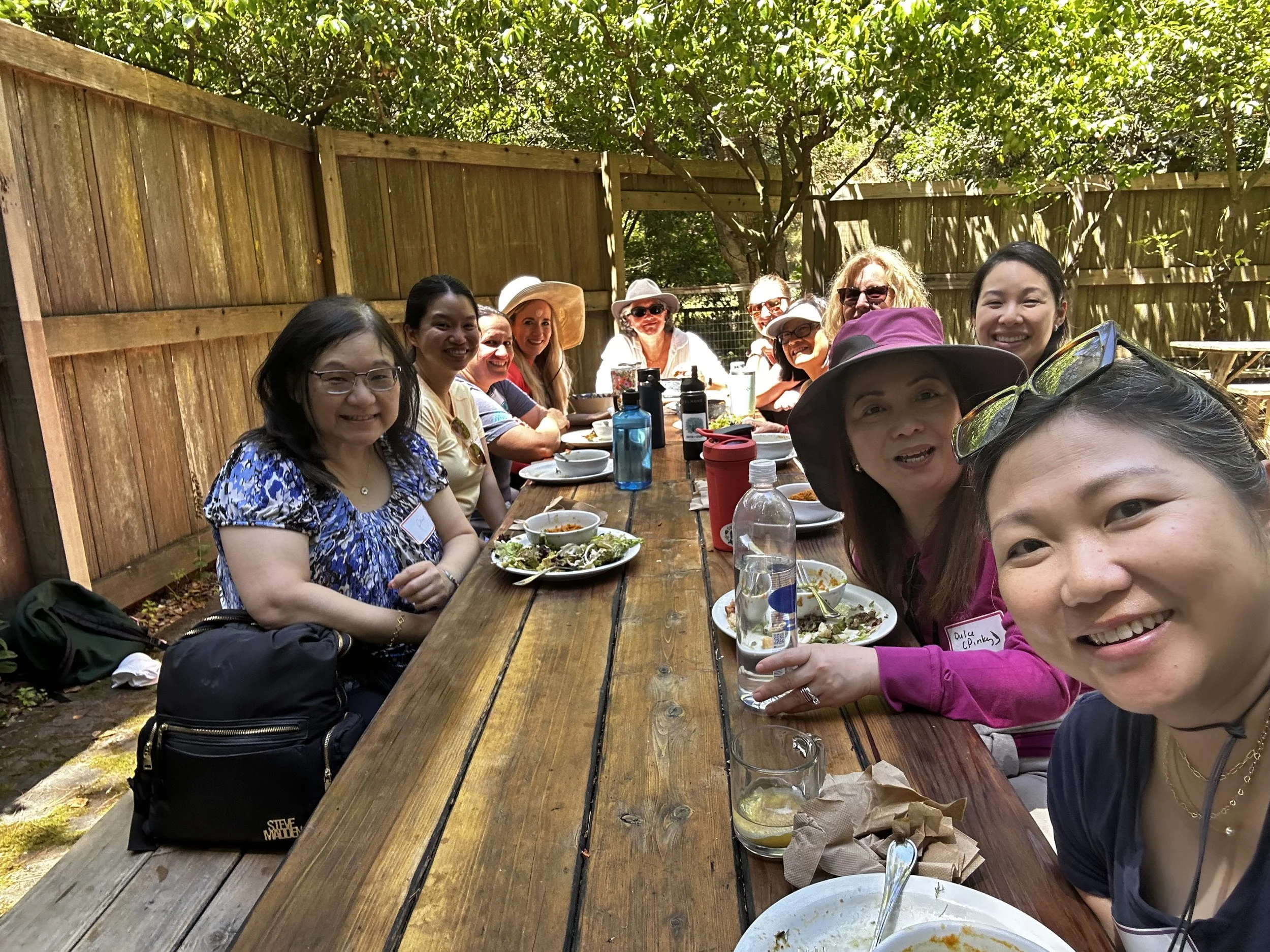 A group of women sitting at a long outdoor wooden table, smiling, with food and drinks. There are trees and a wooden fence in the background.