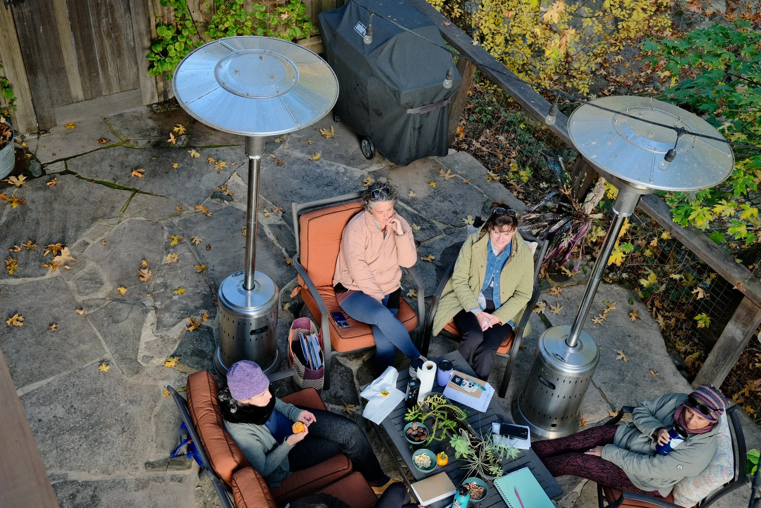 Four women sitting around a table on an outdoor patio with plants and fallen leaves, two heaters, and a barbecue grill in the background, engaged in conversation and eating snacks.