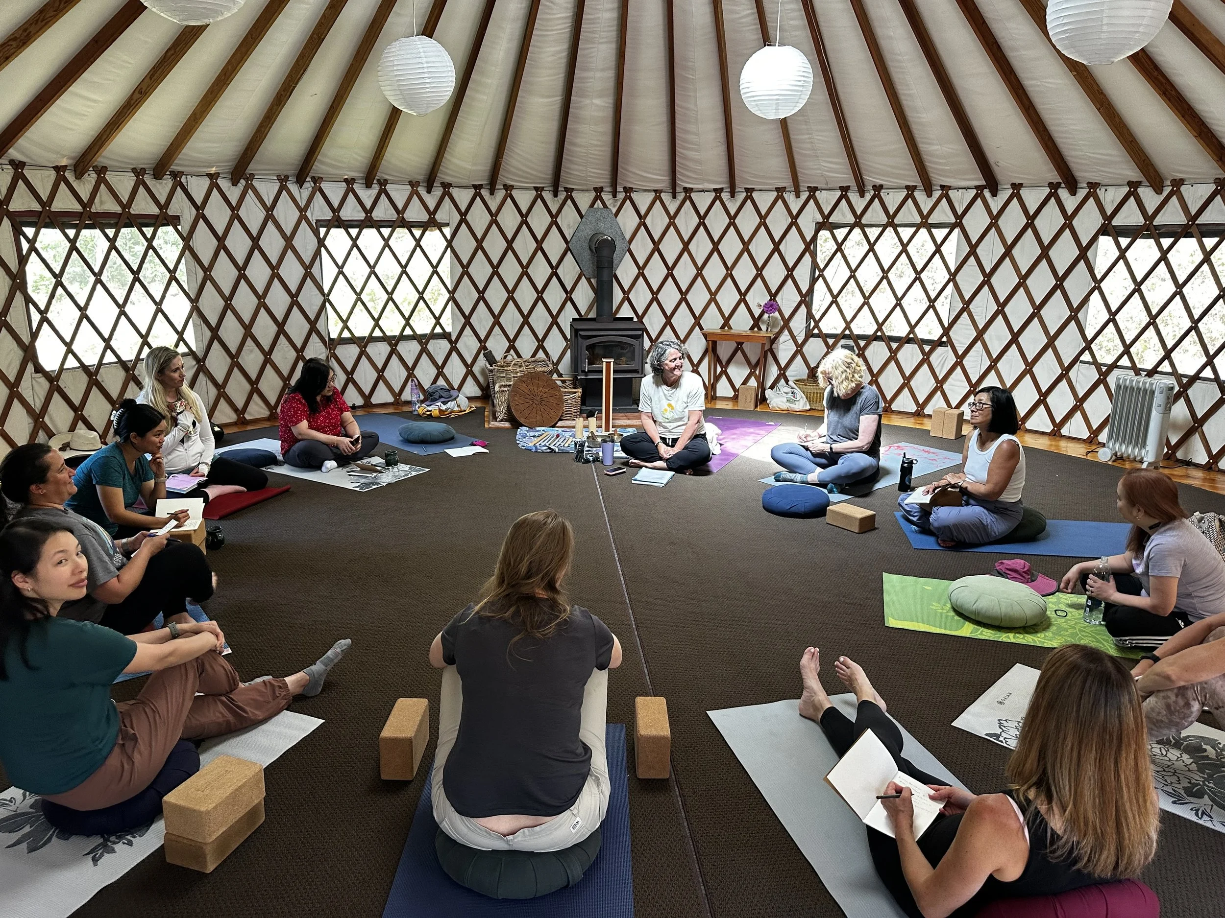 Group of people sitting on yoga mats and blocks in a yurt-like structure with paper lantern lighting, listening to a woman speaking.