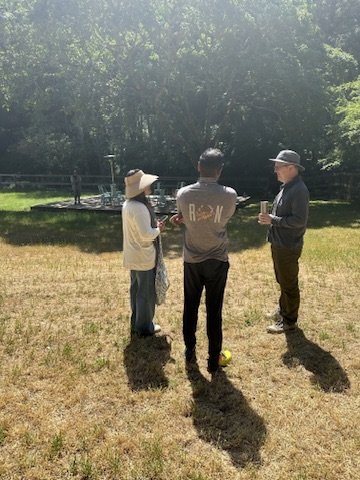 Three people standing on a grassy field next to a pond, engaged in conversation.
