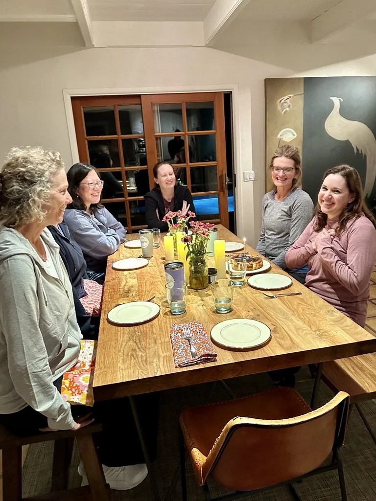 Six women sitting around a wooden dining table, smiling and chatting, with plates, glasses, and a floral centerpiece, in a cozy dining room.