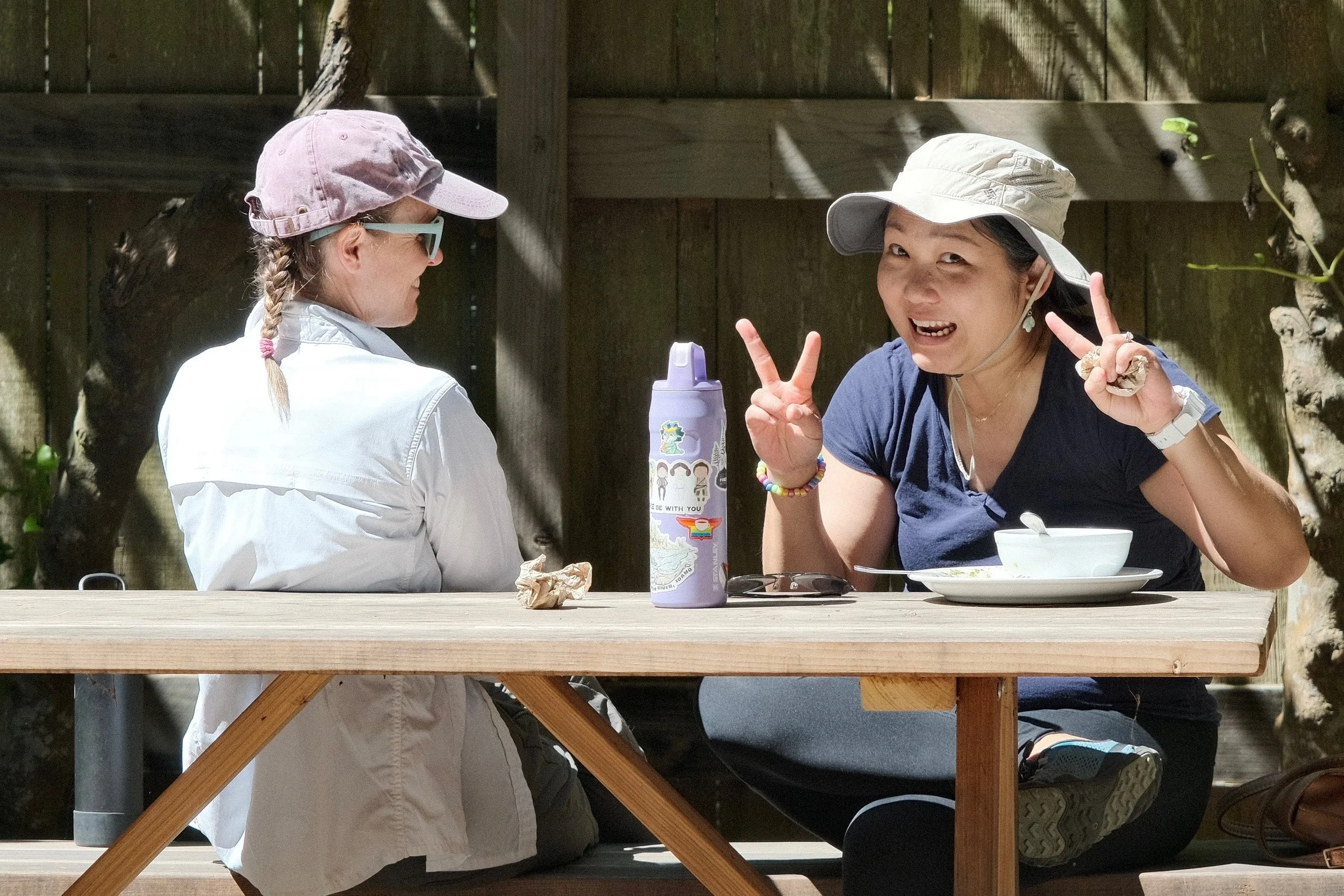 Two women at a picnic table outdoors, one eating and making peace signs, the other facing away wearing a pink hat and sunglasses.