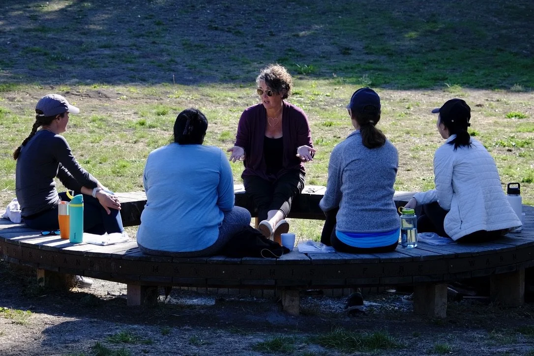 A woman leading a discussion with five women sitting outdoors on a circular wooden bench in a park.