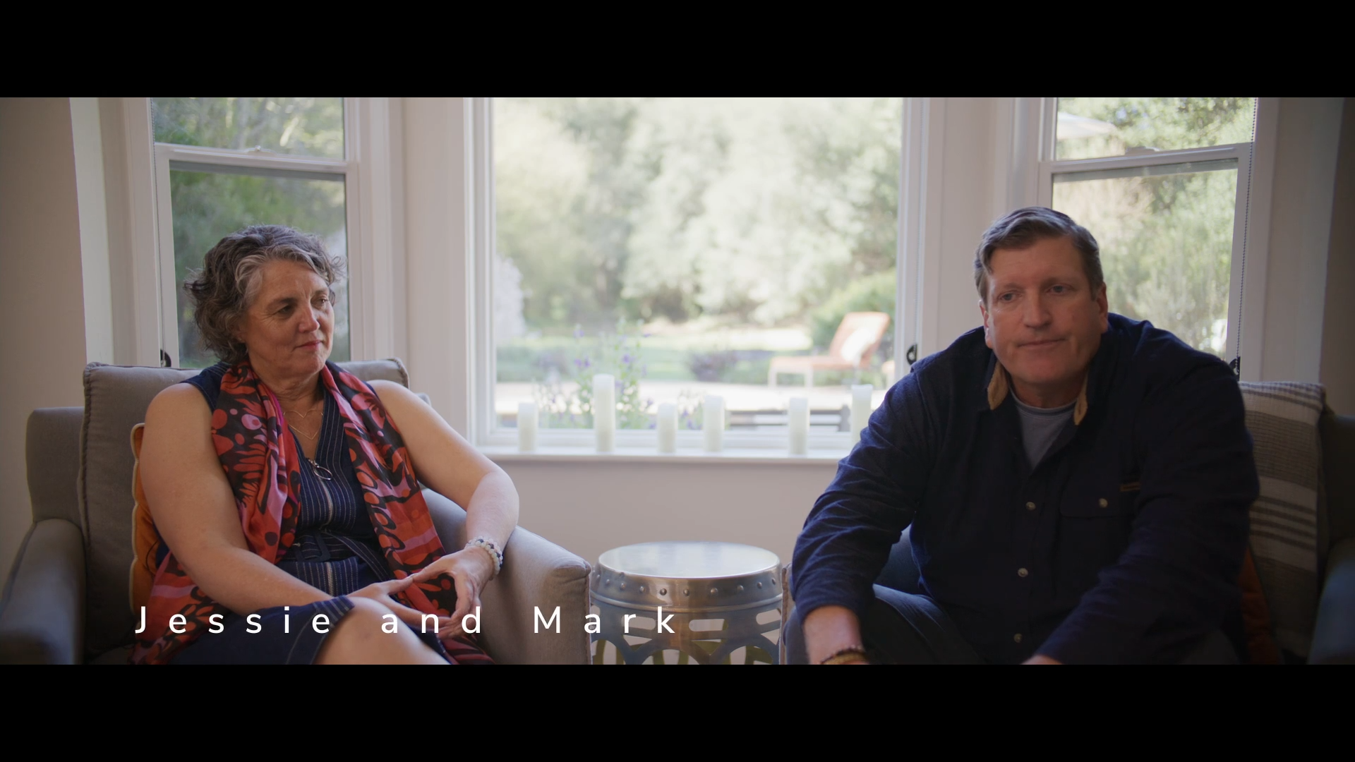 A woman with curly gray hair wearing a sleeveless dress and red scarf sitting on a beige couch, and a man with short brown hair wearing a dark jacket sitting on a chair, both in a living room with large windows showing a garden outside.