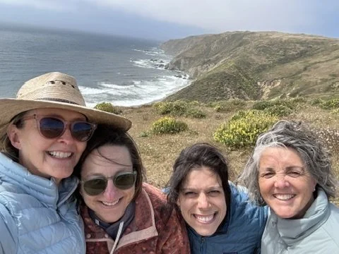 Four women smiling and taking a selfie outdoors with a coastline and cliffs in the background.