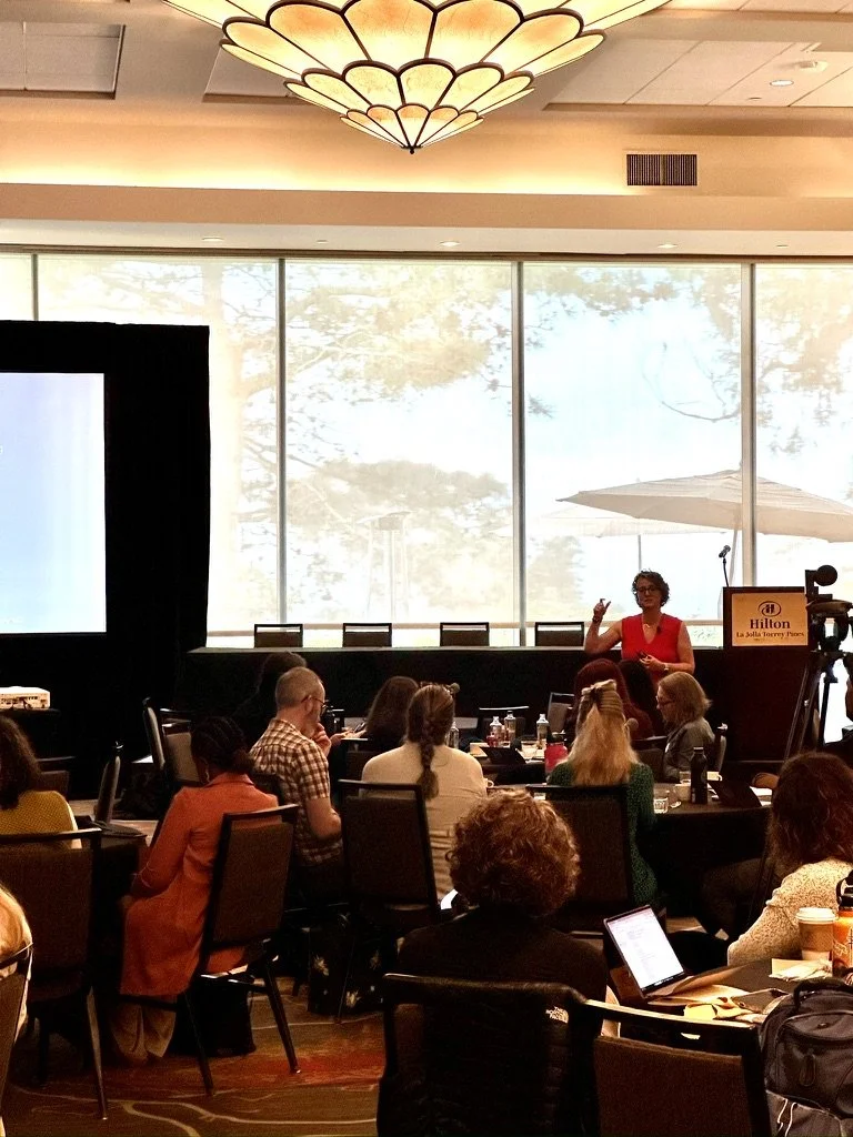 A woman giving a presentation to a group of people seated at round tables in a conference room with large windows and natural light.
