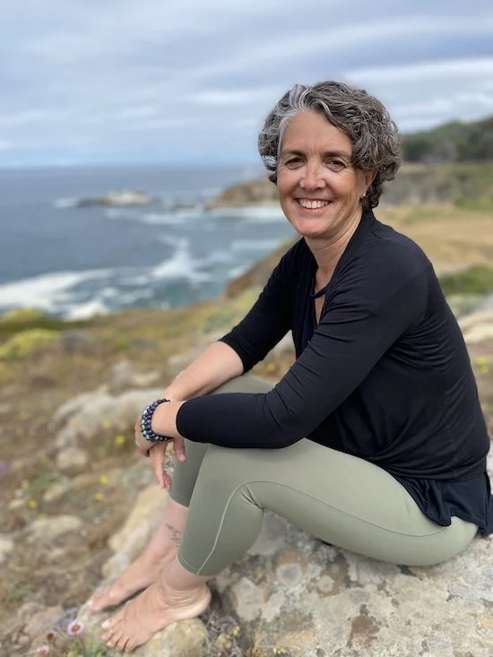 A woman with short gray curly hair sitting on rocks near the coast, smiling, wearing a black long-sleeve shirt and beige leggings, with a view of the ocean and cliffs in the background.