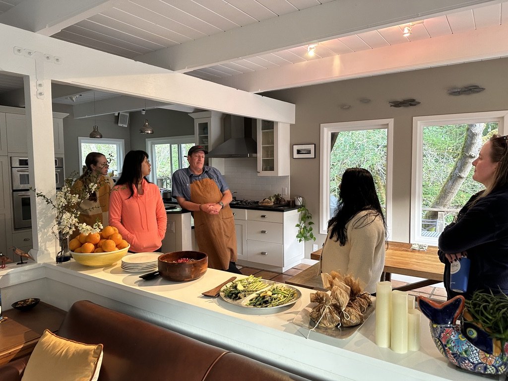Group of five people gathered in a kitchen, with a man in an apron speaking to the women. The kitchen has white cabinets, windows showing trees outside, and a countertop with plates, a bowl of oranges, and wrapped items.