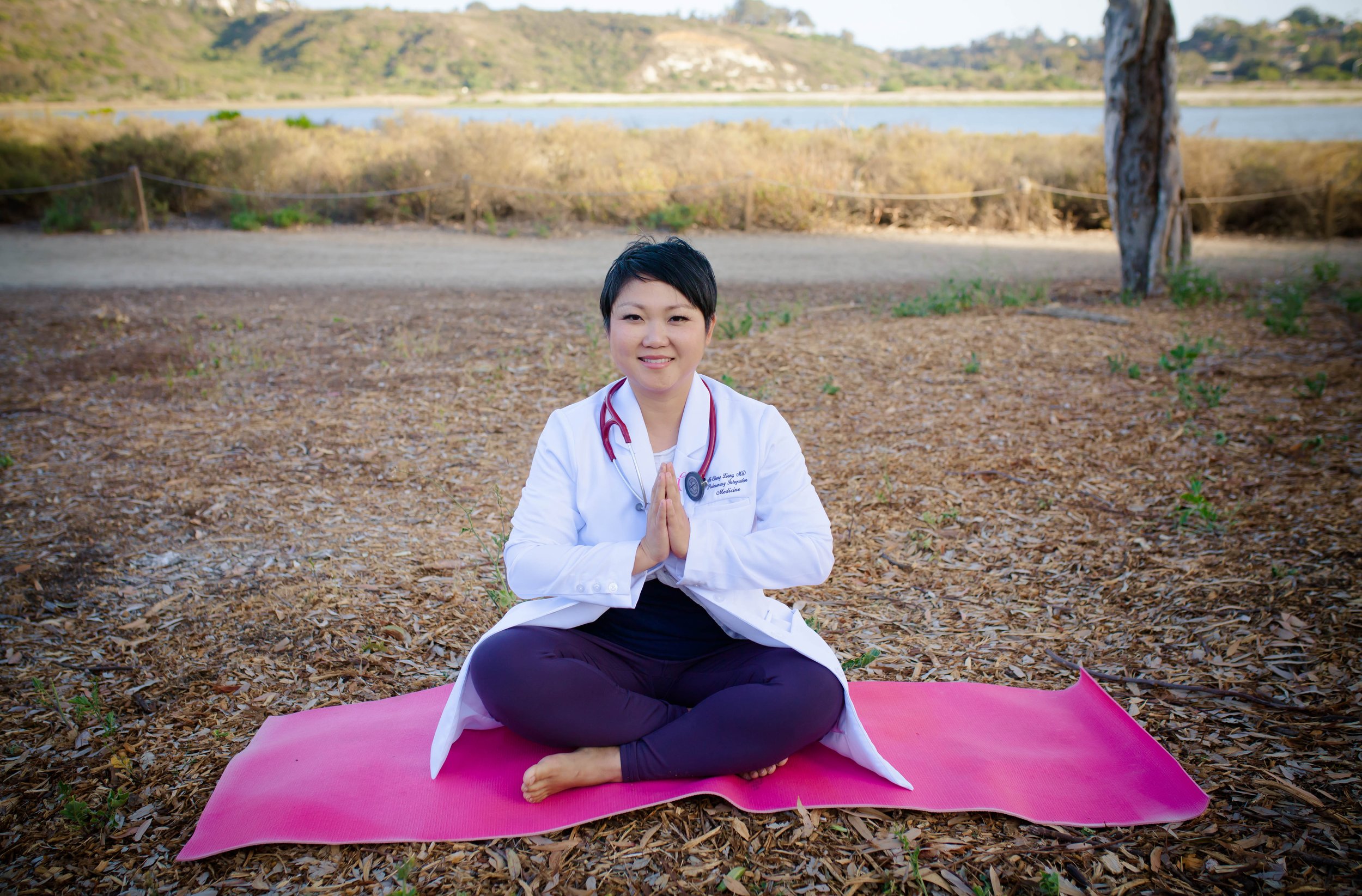 A woman dressed as a doctor practicing yoga outdoors on a pink mat, sitting cross-legged with hands in a prayer position, in a natural setting near a body of water under a tree.