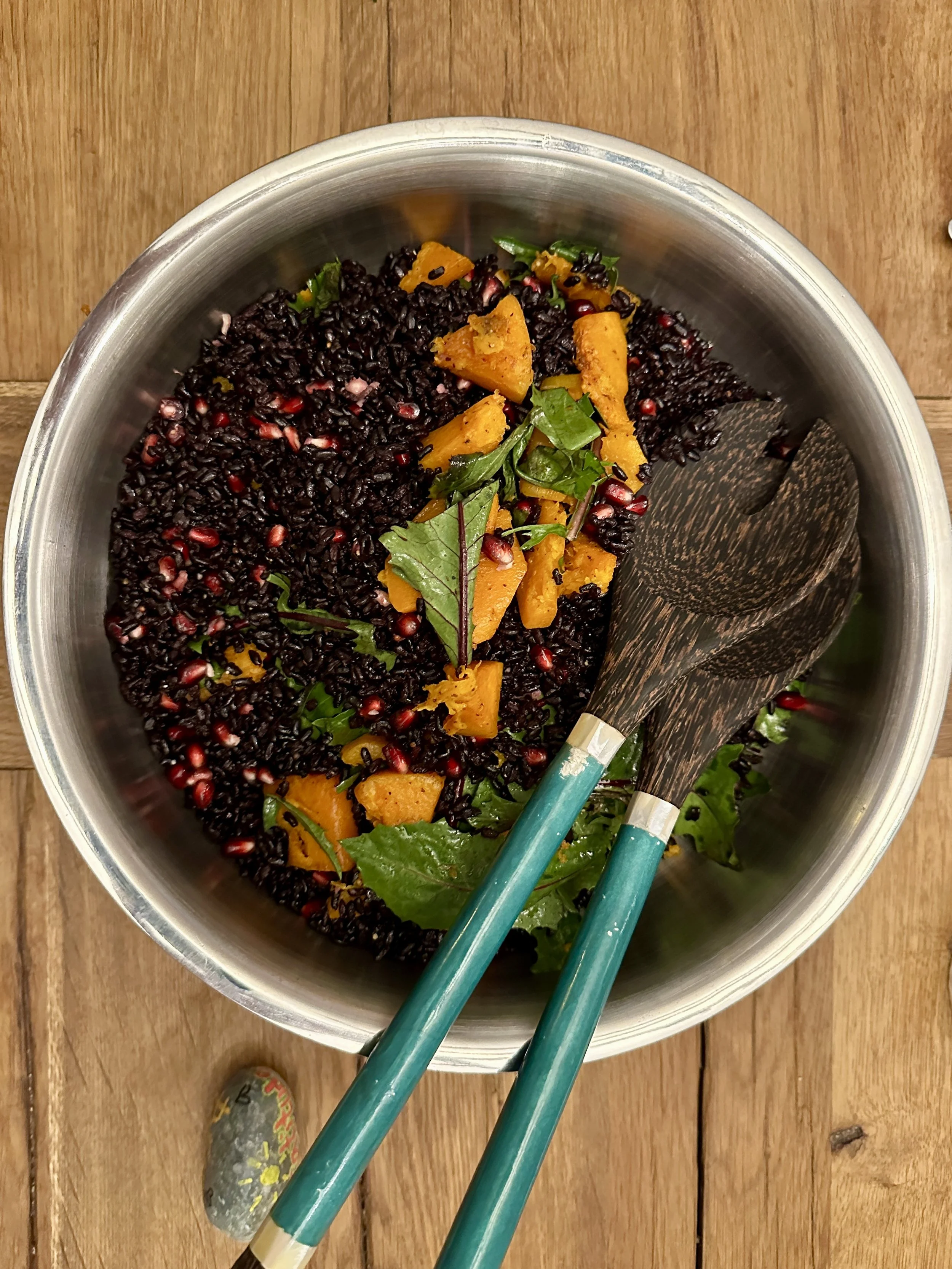 A bowl of cooked black rice with pomegranate seeds, roasted pumpkin pieces, and green leafy herbs, placed on a wooden surface with a pair of black and teal chopsticks inside the bowl.
