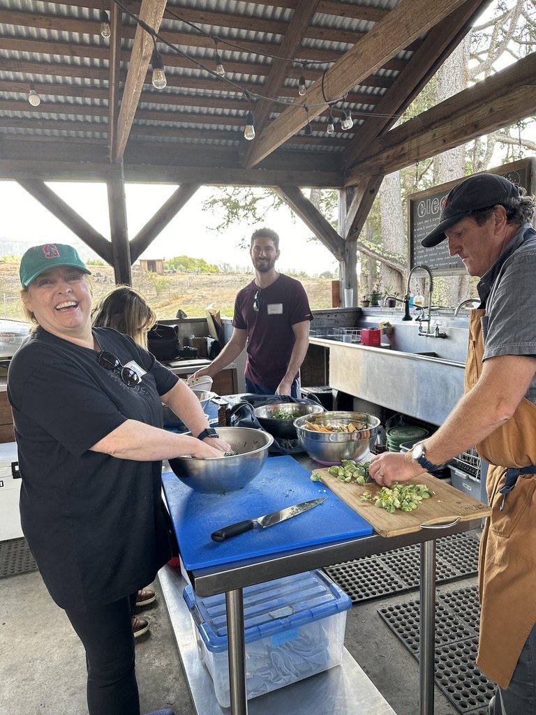 Three people cooking outdoors under a wooden shelter, with a woman smiling and a man chopping vegetables.