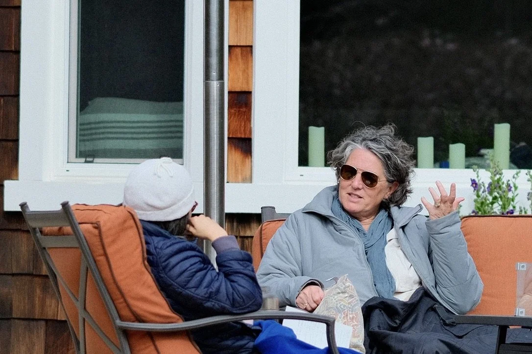 An older woman with sunglasses talking to a younger person in a white hat, both sitting outdoors on orange cushioned chairs.