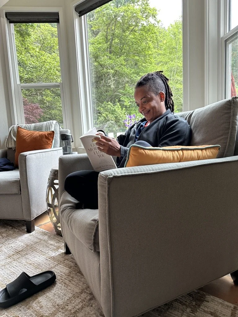 A person with dreadlocks is sitting on a beige armchair, reading a book in a bright living room with large windows showing green trees outside.