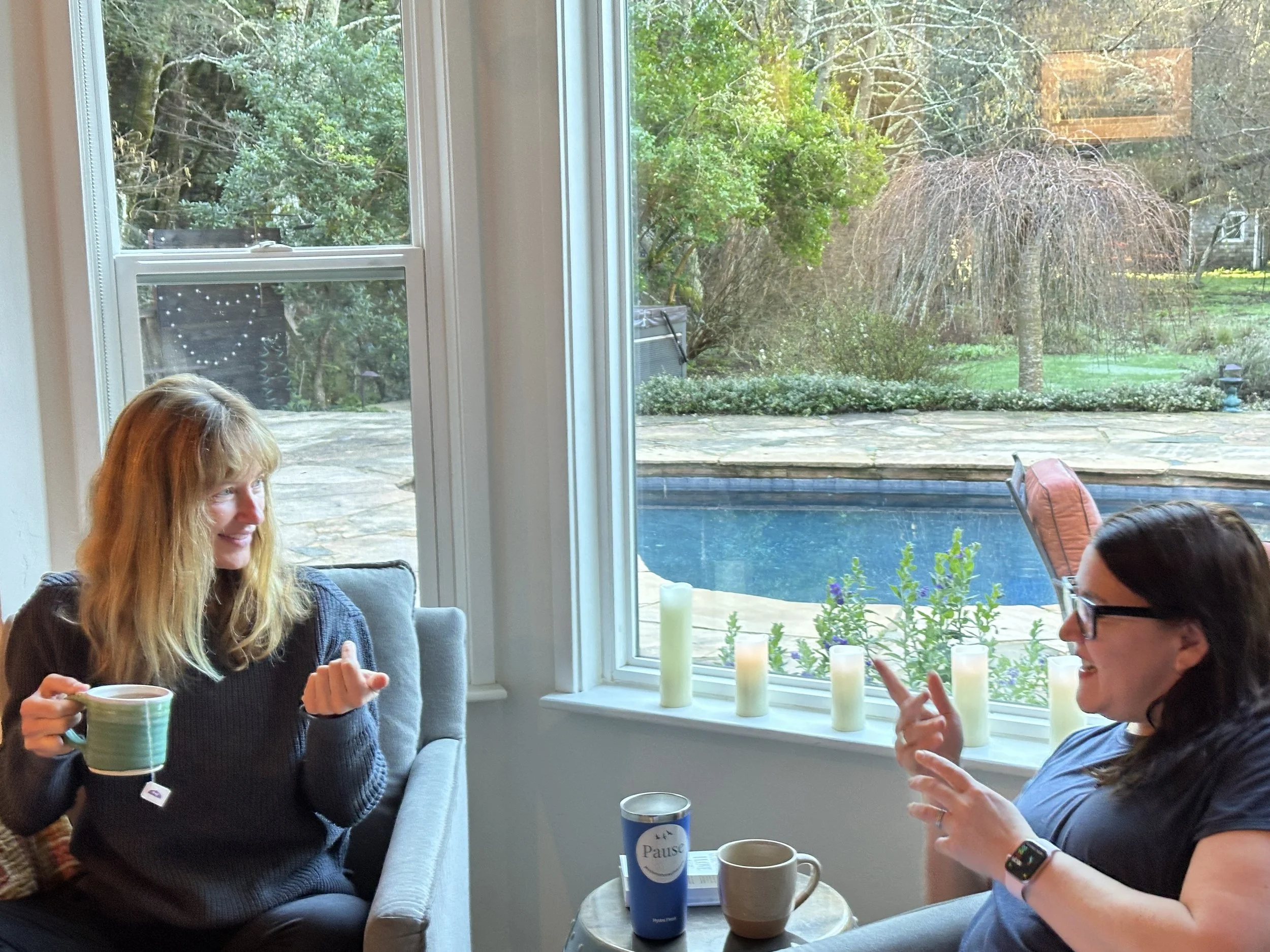 Two women sitting in a cozy living room by a large window, engaging in conversation with mugs in hand, overlooking a backyard with a swimming pool and trees.