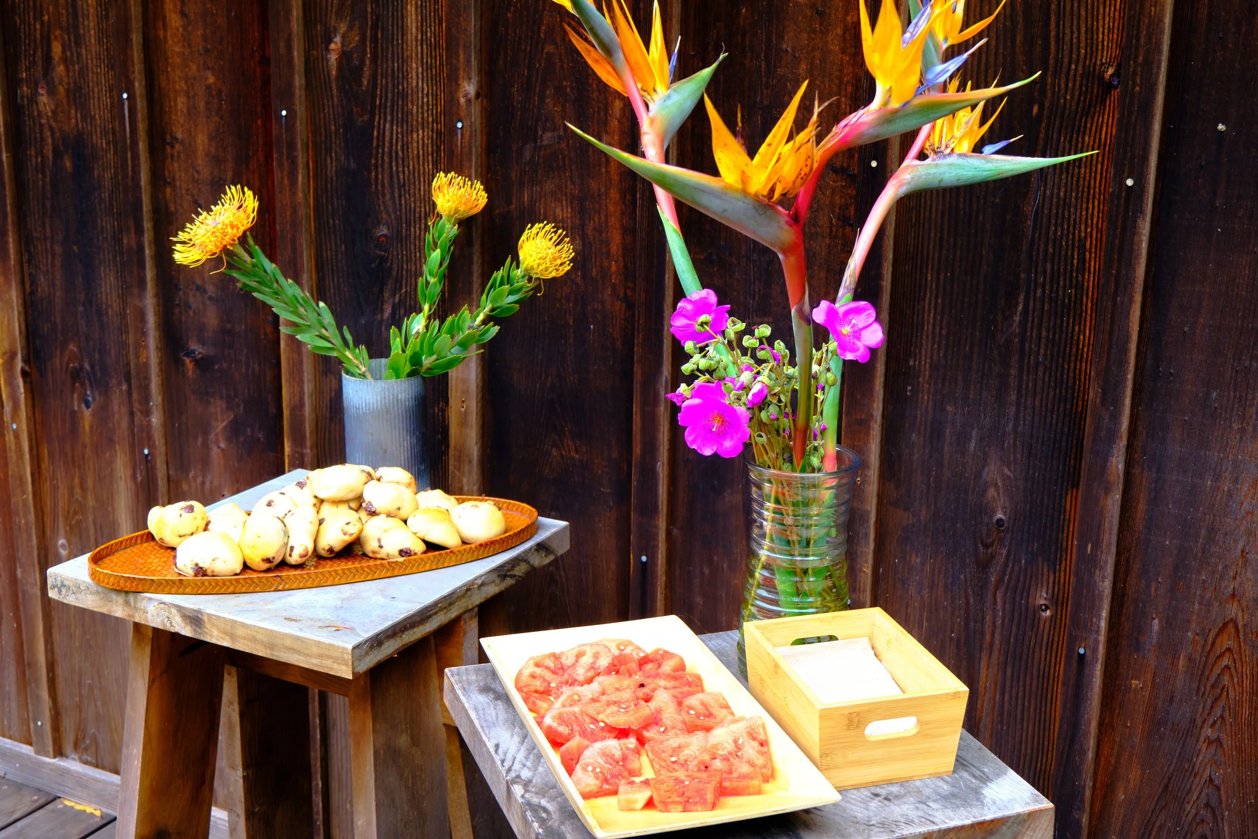 Outdoor display of tropical fruits and flowers against a wooden fence. There is a tray of sliced watermelon, a plate of scones or pastries, and vases with colorful flowers including heliconias, pink flowers, and yellow and green foliage.