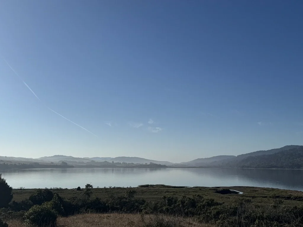 A peaceful lake surrounded by rolling hills and clear blue sky with wispy clouds and contrails.