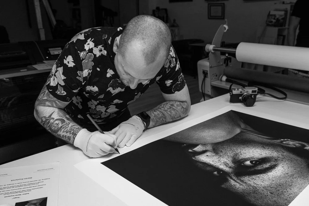 A man with tattoos on his arms and wearing gloves is signing a large black-and-white portrait of a woman's face on a table.