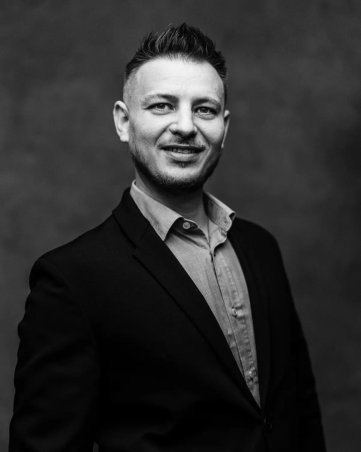 Čiernobiely portrét muža v obleku a košeli, ktorý sa usmieva do kamery.